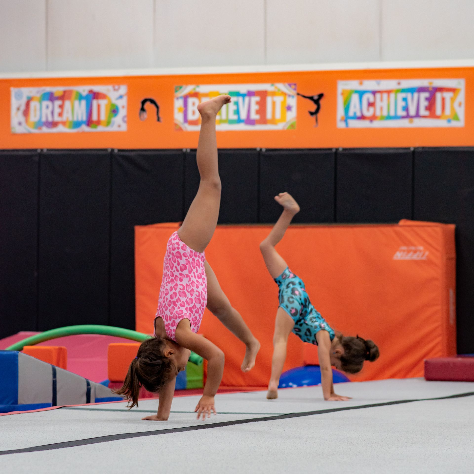 Two gymnasts doing handstands on a mat beneath “Dream It, Believe It, Achieve It” banners.