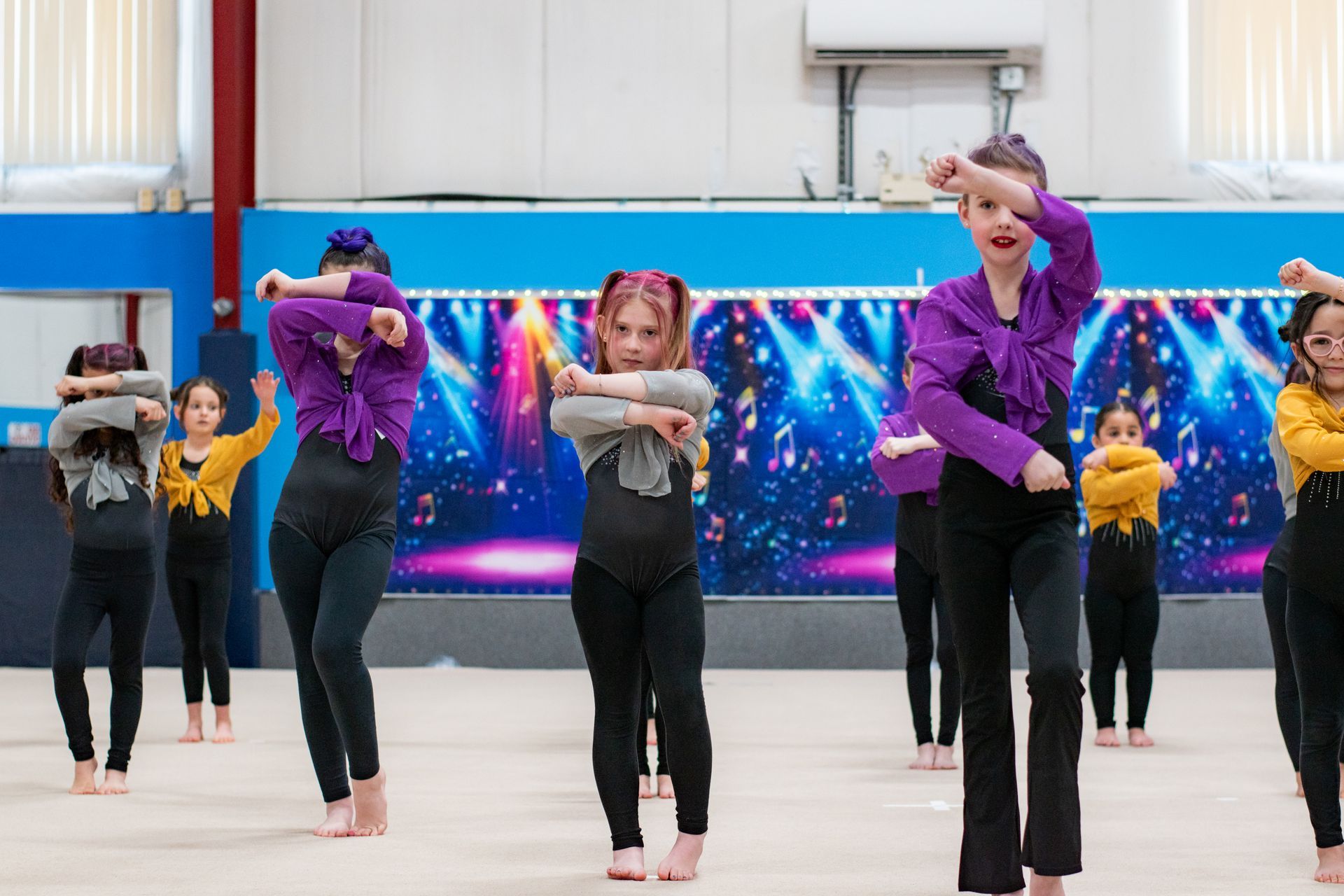Dancers rehearsing in a studio, with three women in purple and gray at front and others in yellow behind.