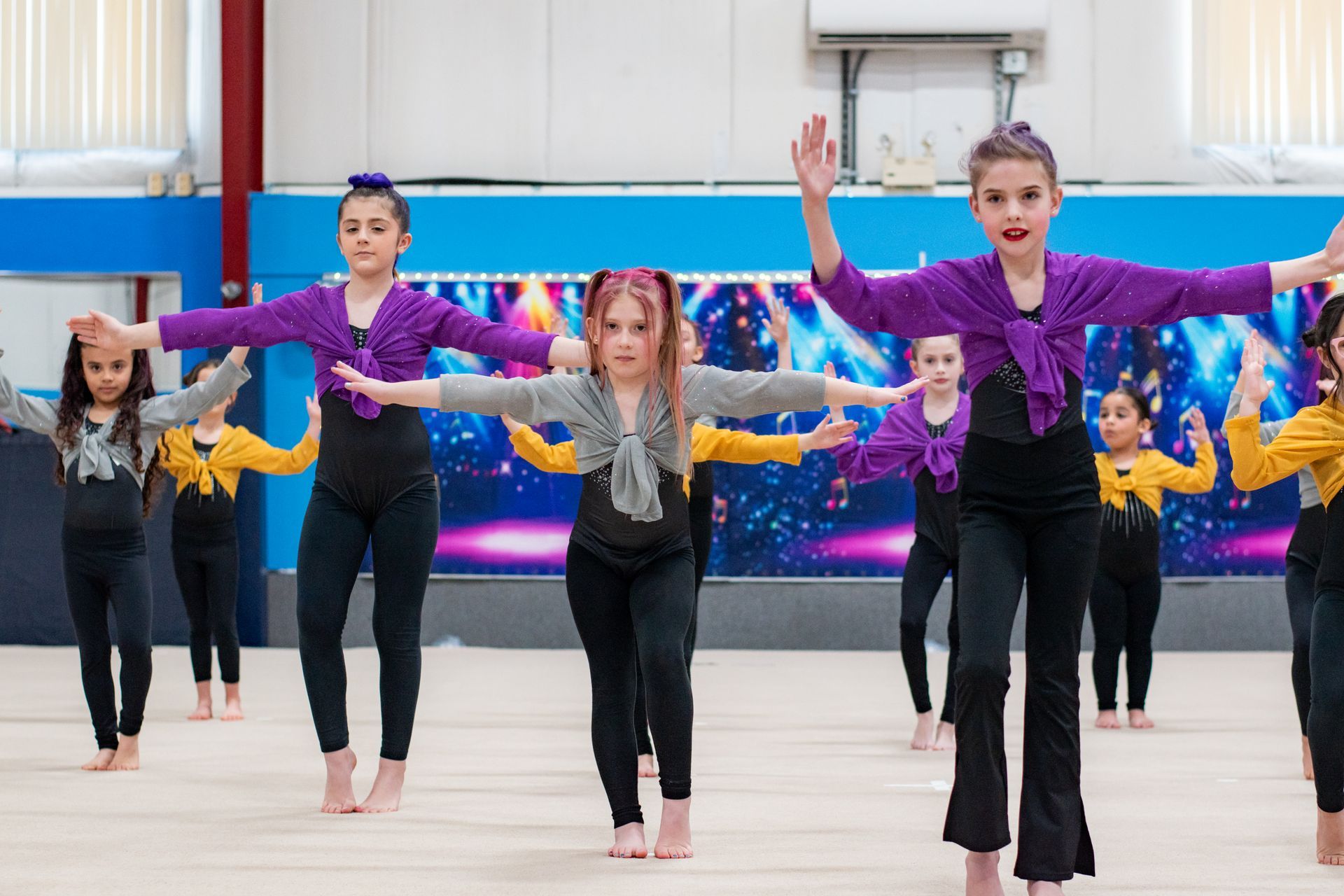 Children dancing in purple and gold costumes in a gymnasium, arms raised in formation.