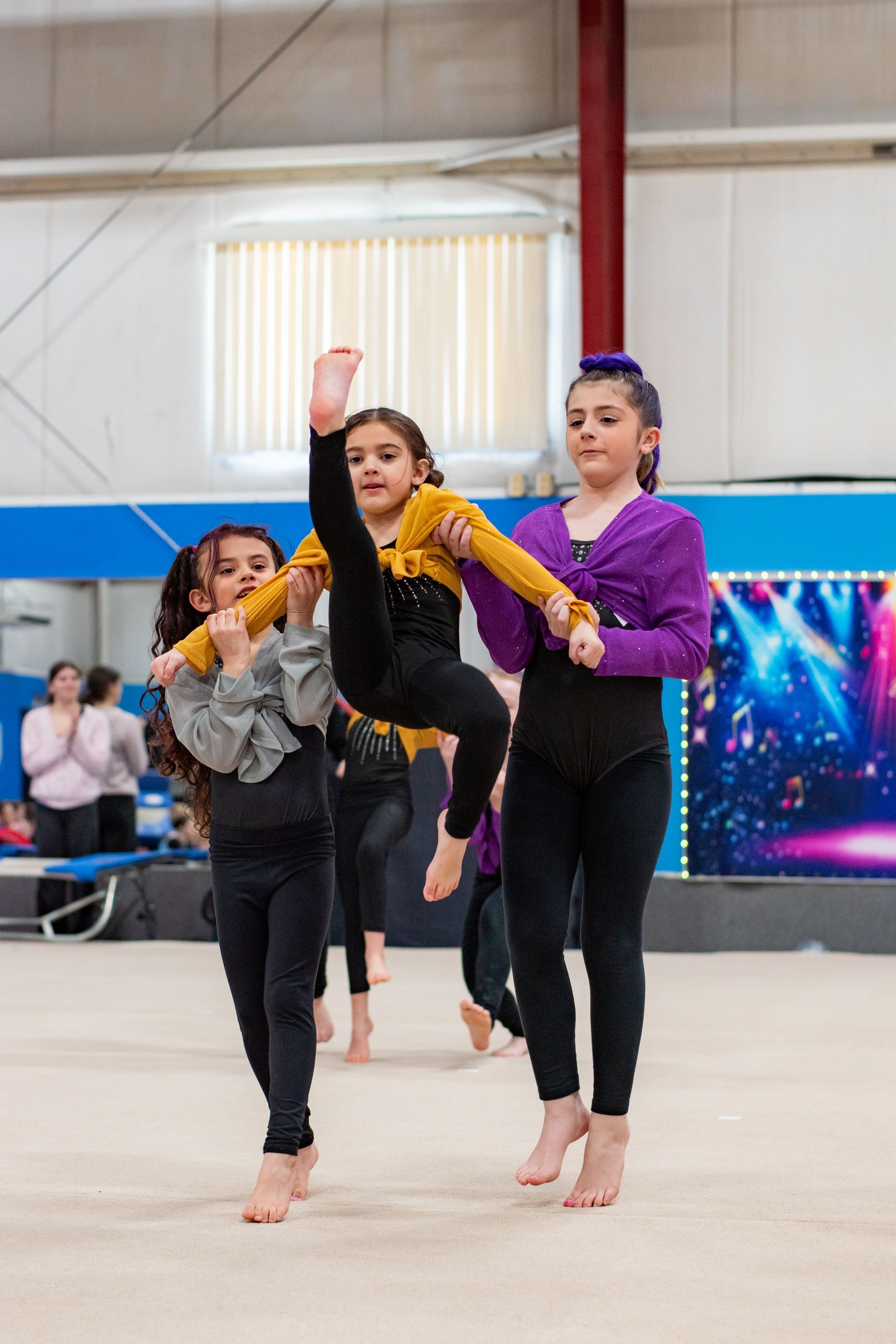 Three gymnasts in colorful leotards perform a balance pose on a gym floor indoors.