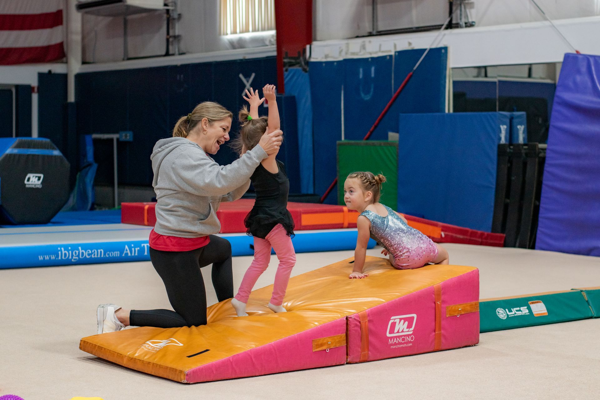 Coach helping two children practice a gymnastics move on an orange mat in a gym