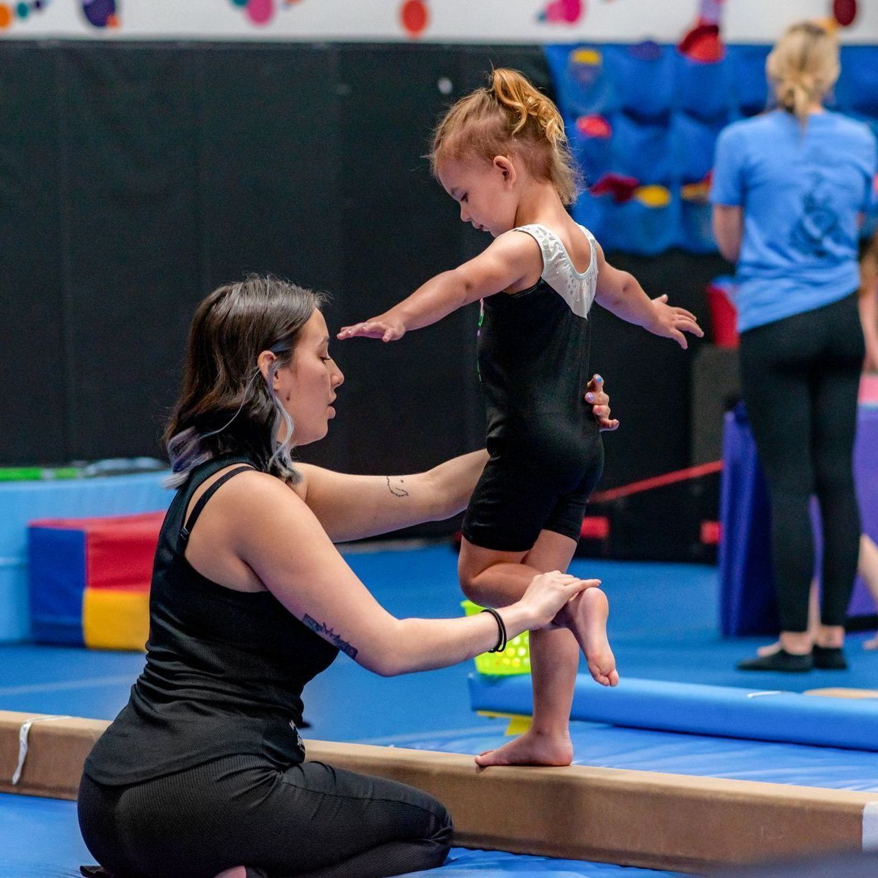 Gym coach spotting a young gymnast balancing on a beam in a colorful gymnasium