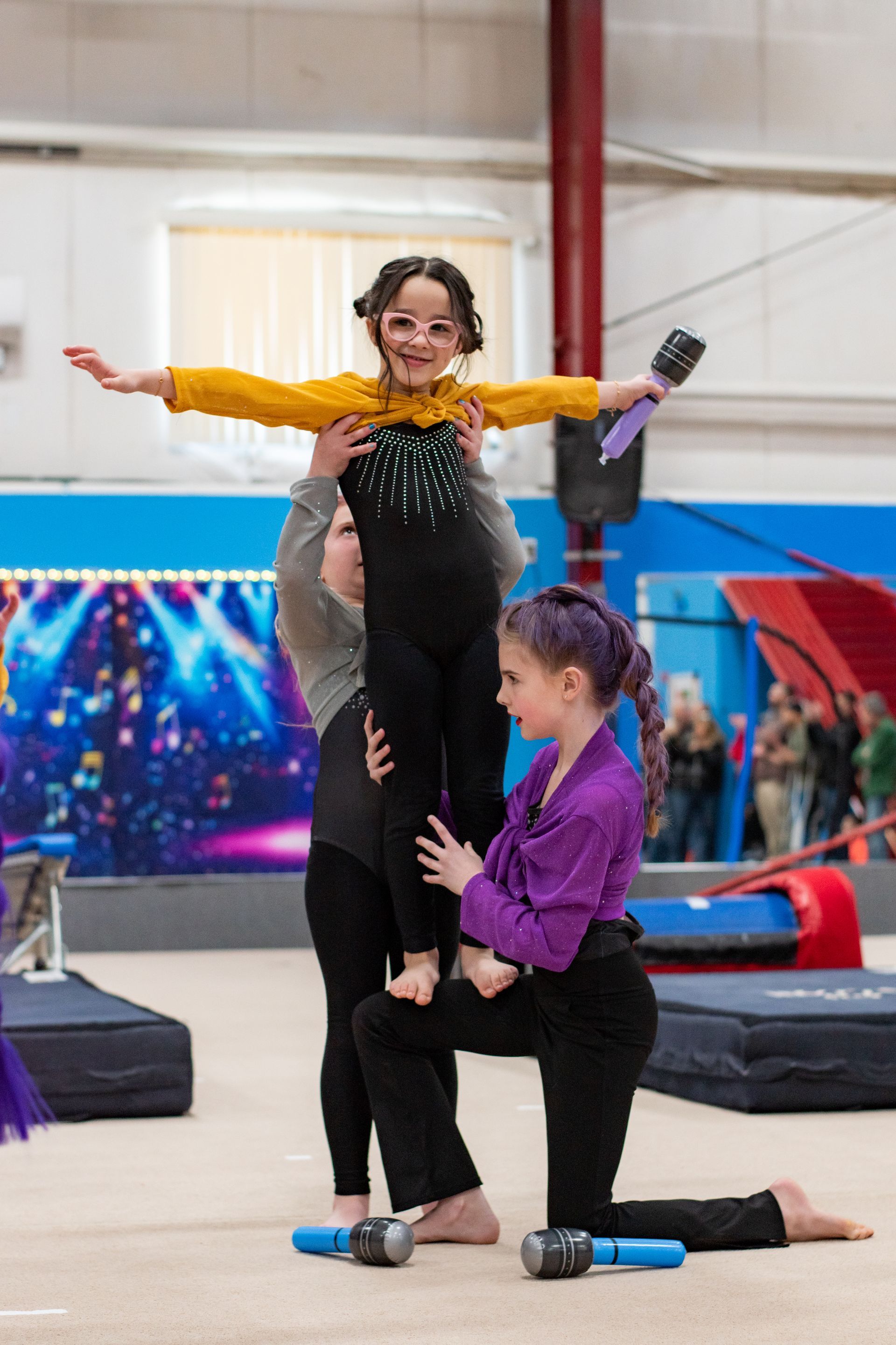 Gymnast balancing on two assistants in a gym, arms outstretched, with colorful mats and banners behind.