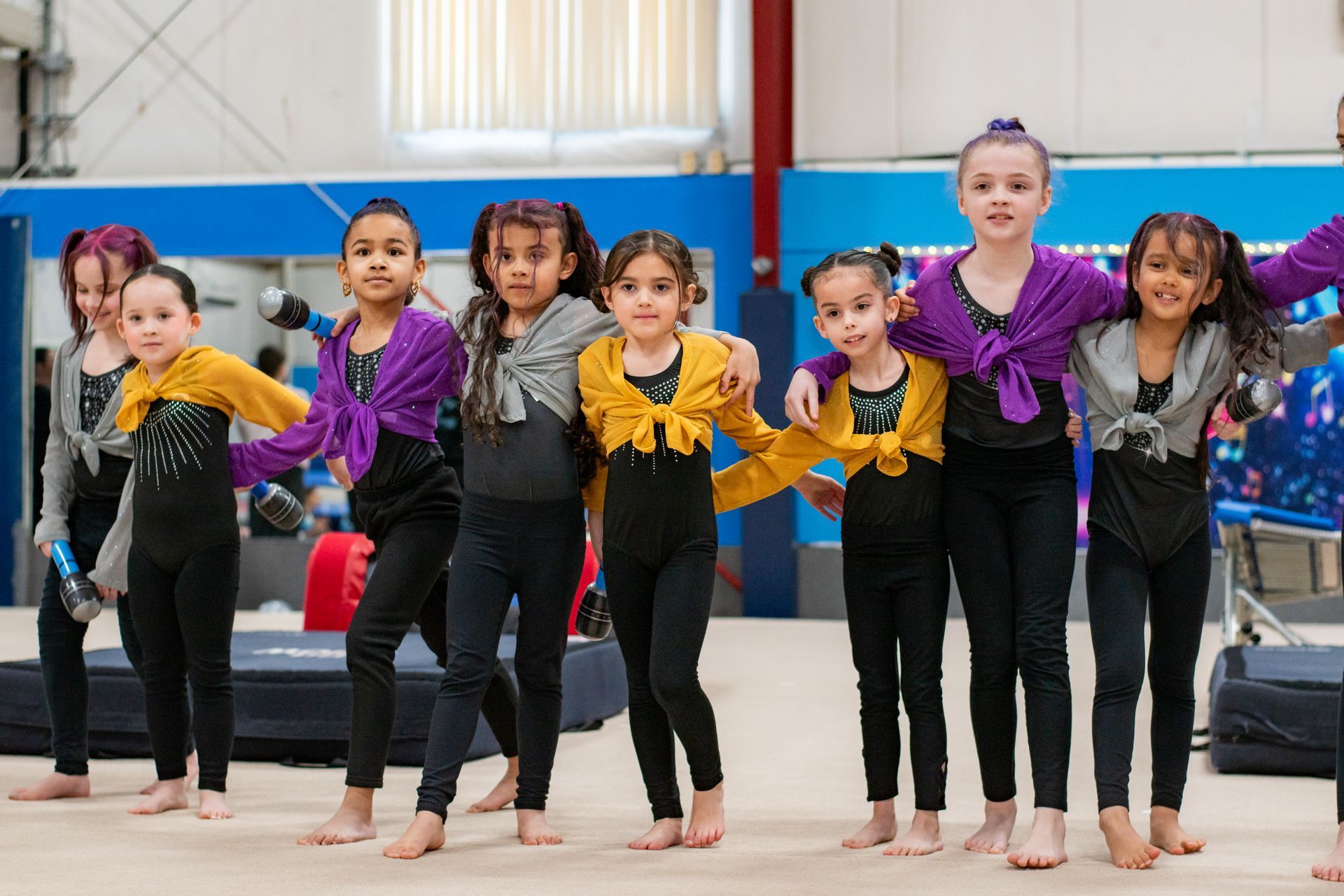 Six children in colorful leotards pose on a gym floor with arms raised in a dance routine.