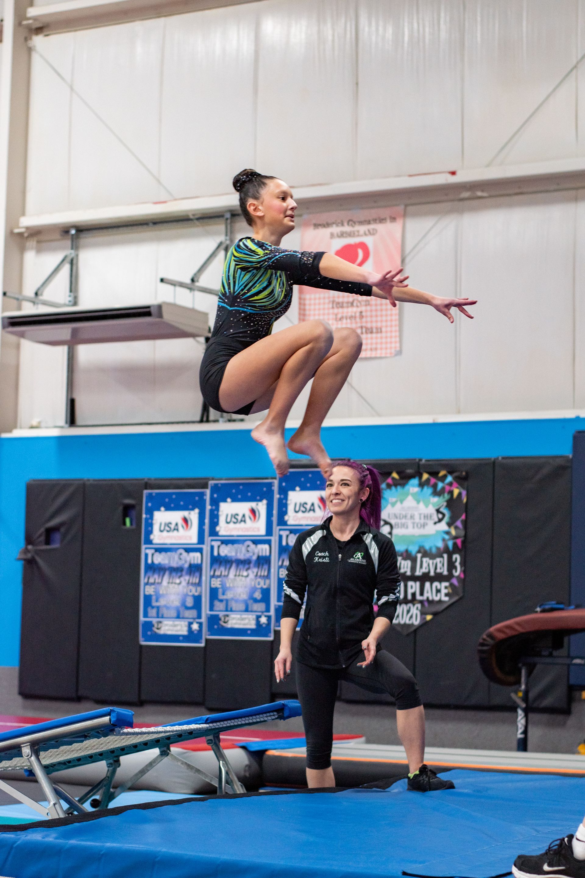 Gymnast midair over a trampoline, with a coach watching in an indoor gym.