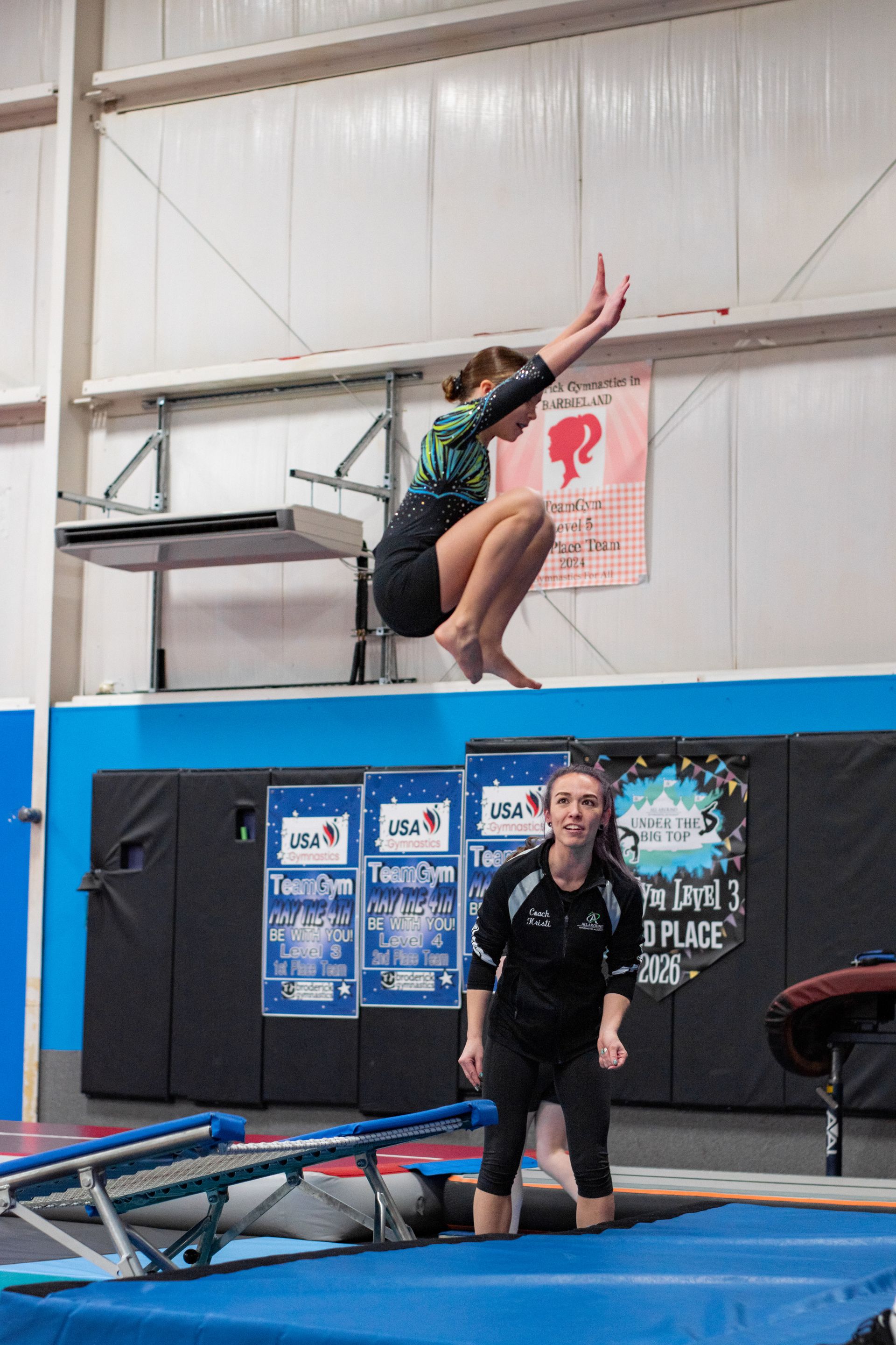 Gymnast midair on a trampoline indoors, with coach watching nearby.