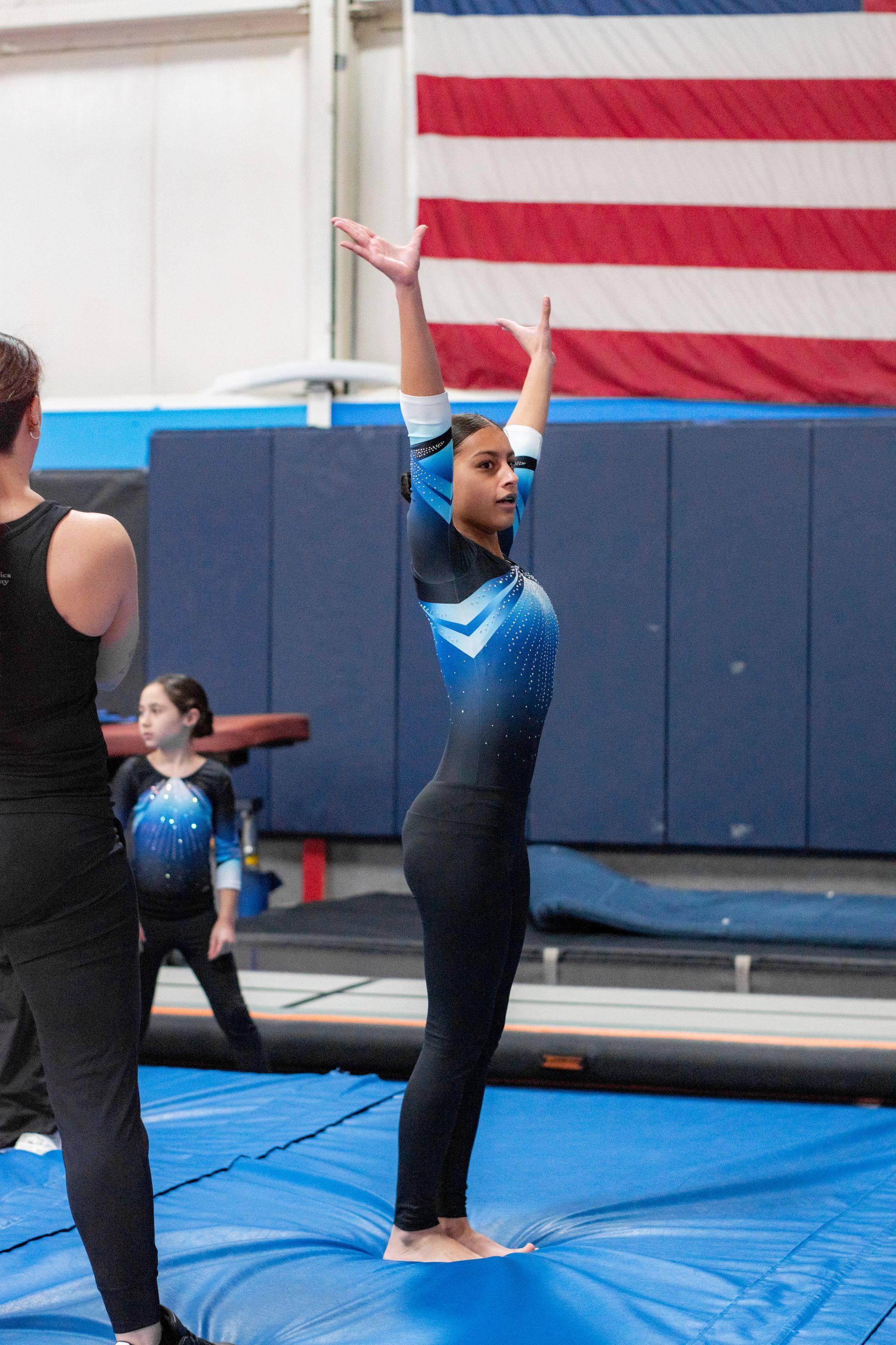 Gymnast in blue leotard posing on a mat in a gymnasium with an American flag behind her.