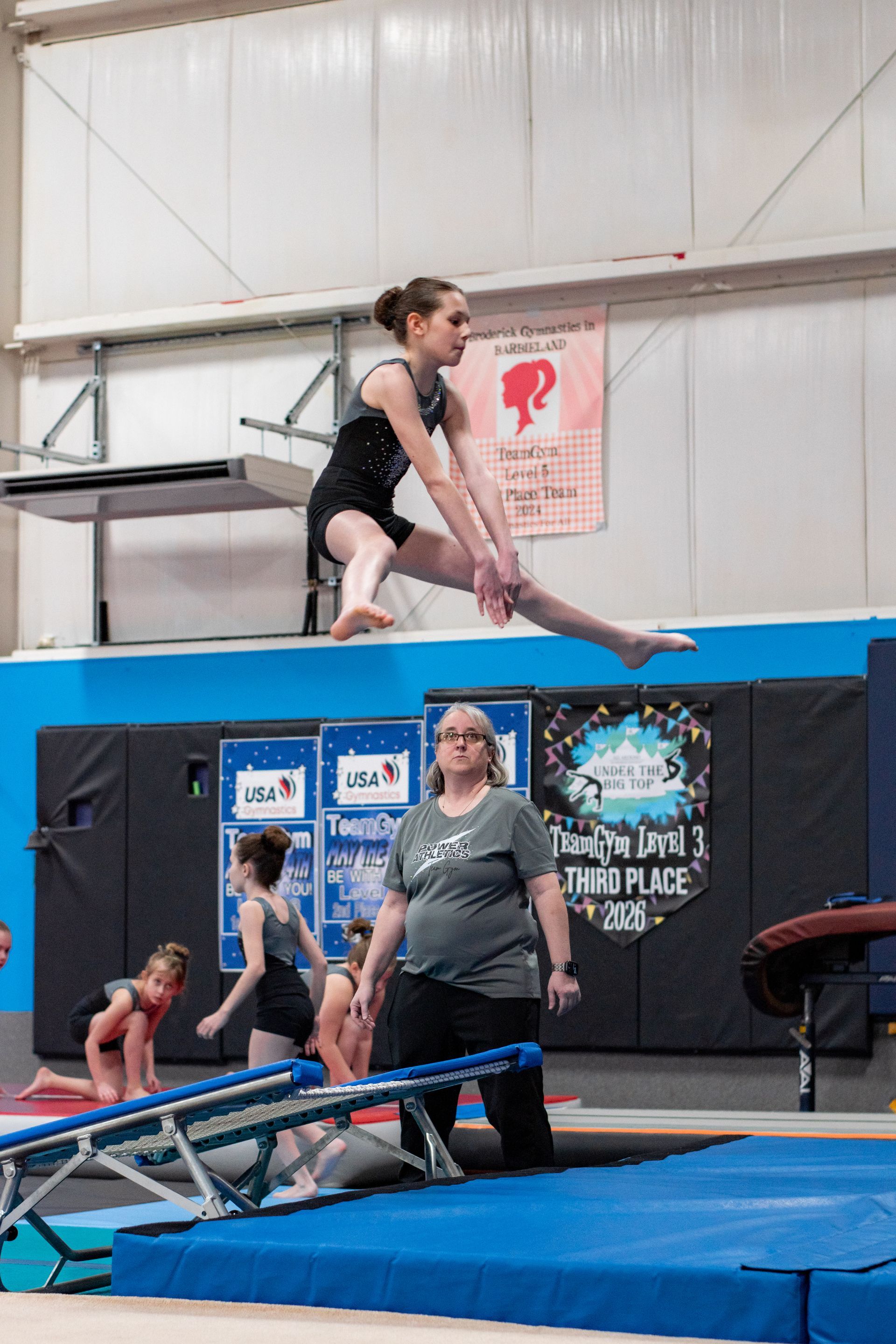 Gymnast balancing on a beam in an indoor gym, with a coach and other athletes below.