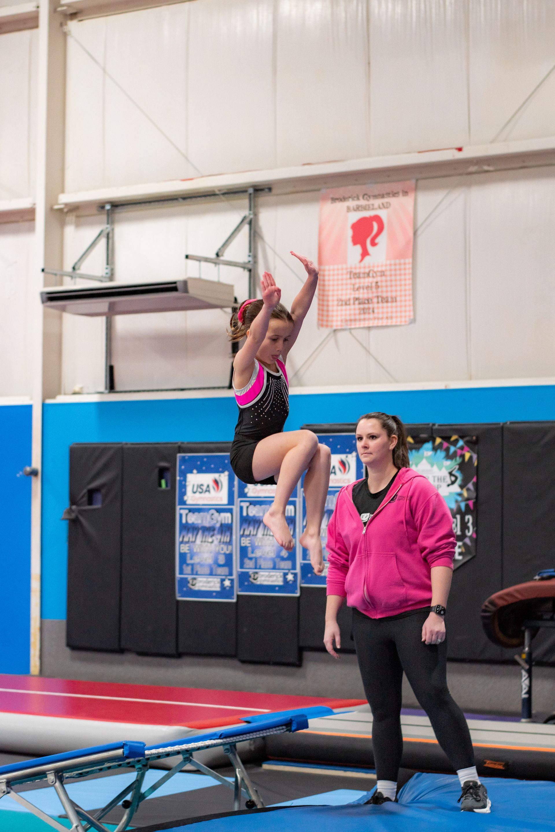 Gymnast hangs from a bar while a coach watches in an indoor gym with mats.