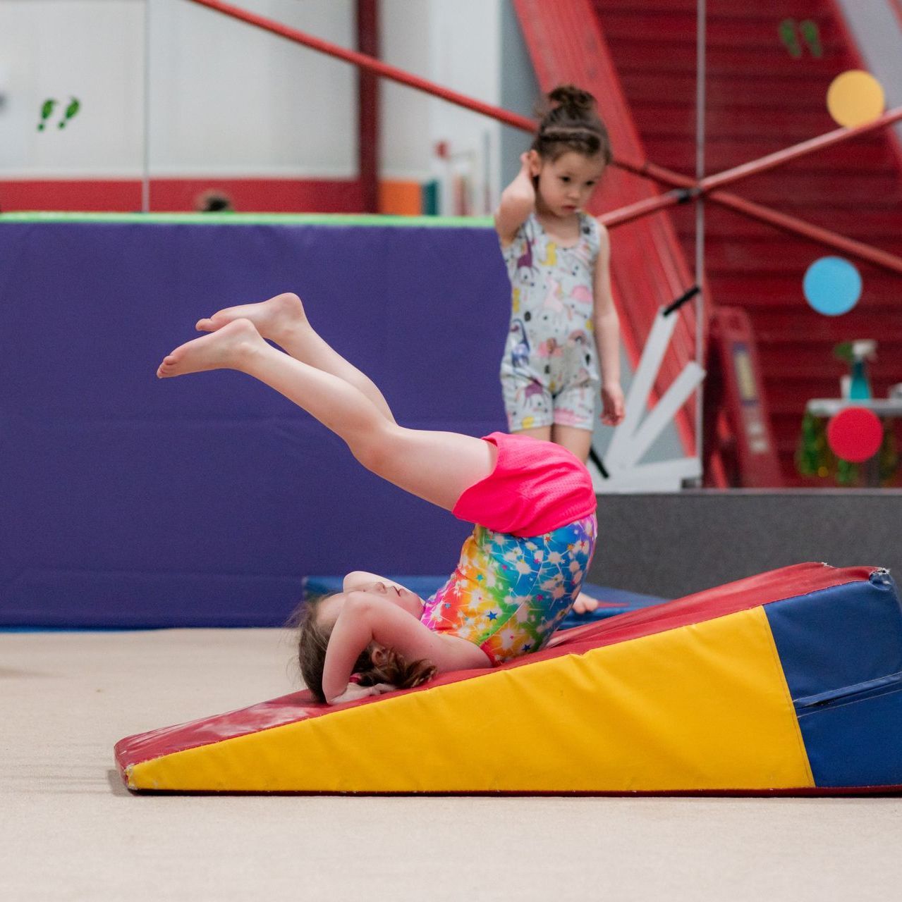 Child doing a backward roll on a colorful foam wedge in a gym while another child watches nearby