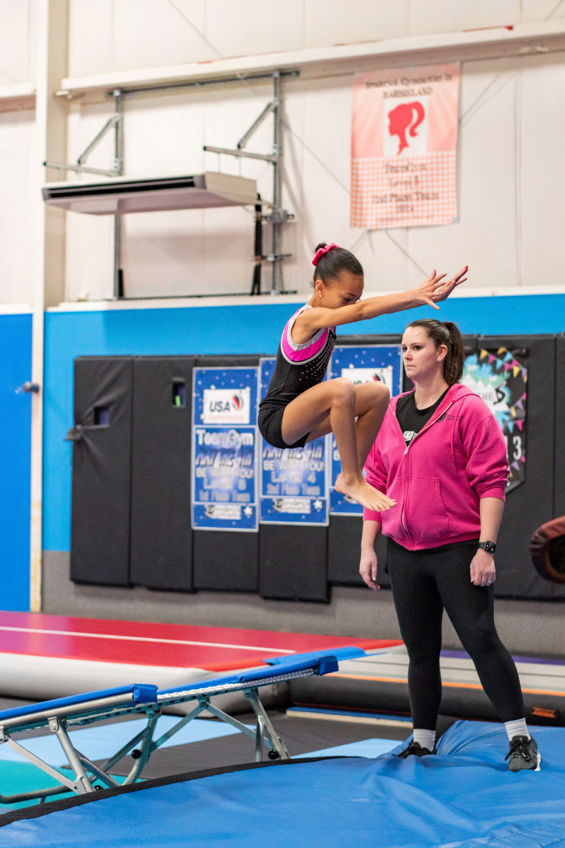 Young gymnast mid-air over a blue trampoline while a coach watches in a gym
