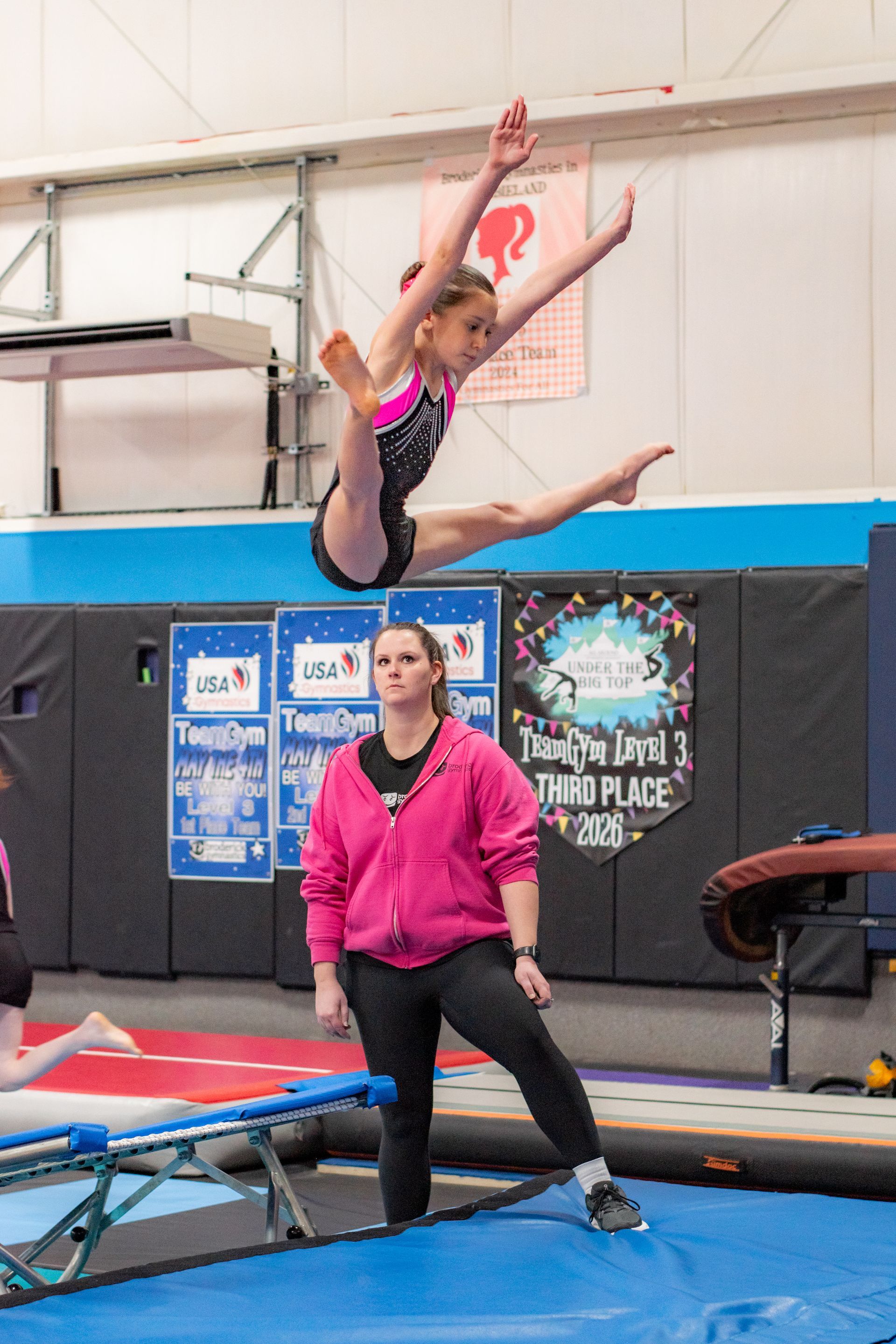 Gymnast mid-flip on an indoor trampoline as a coach watches nearby