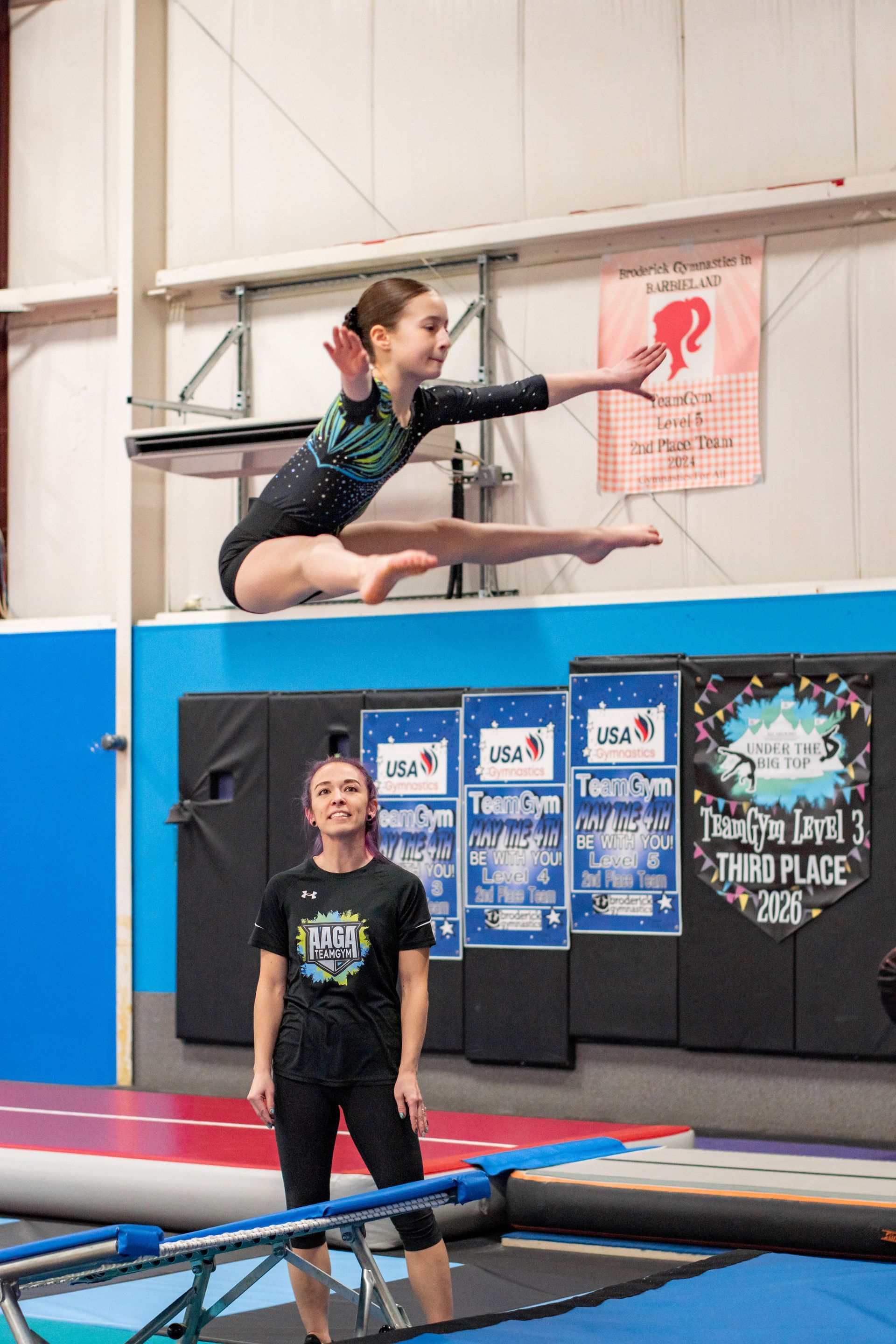Gymnast midair on balance beam while coach stands nearby in an indoor gym