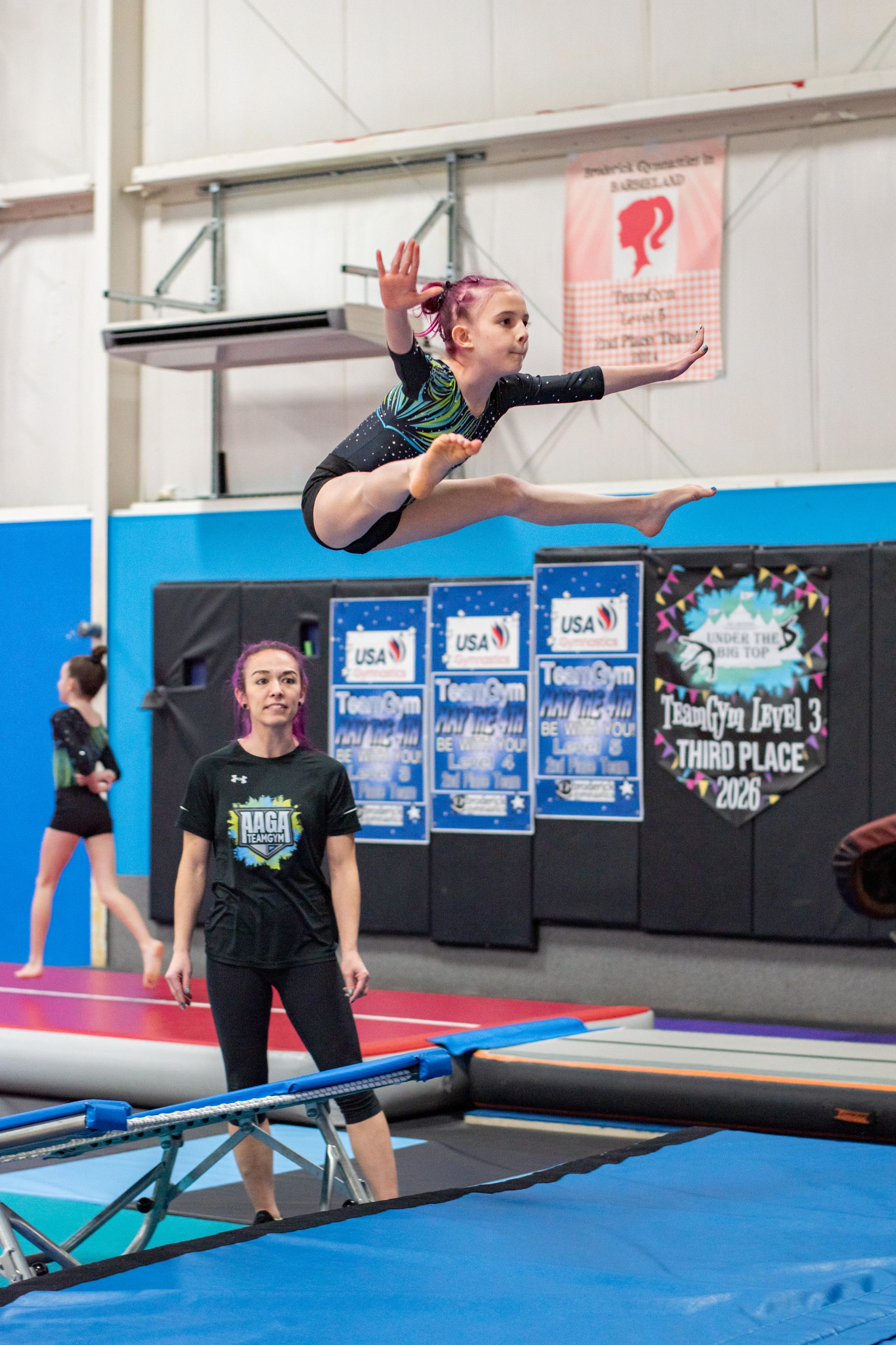 Gymnast balancing on a beam in an indoor gym with blue mats and a trainer behind her