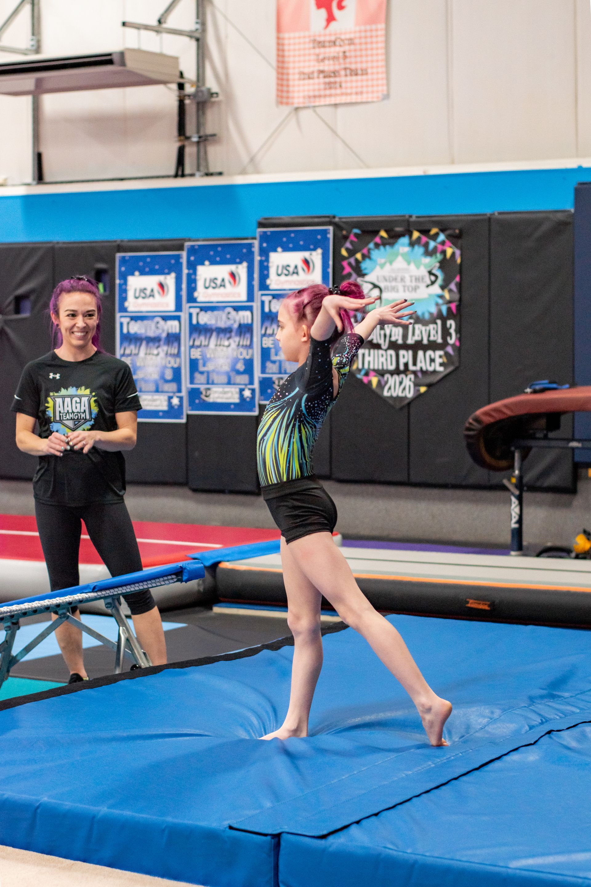 Gymnast balancing on a blue mat in a gym, with a coach standing nearby.