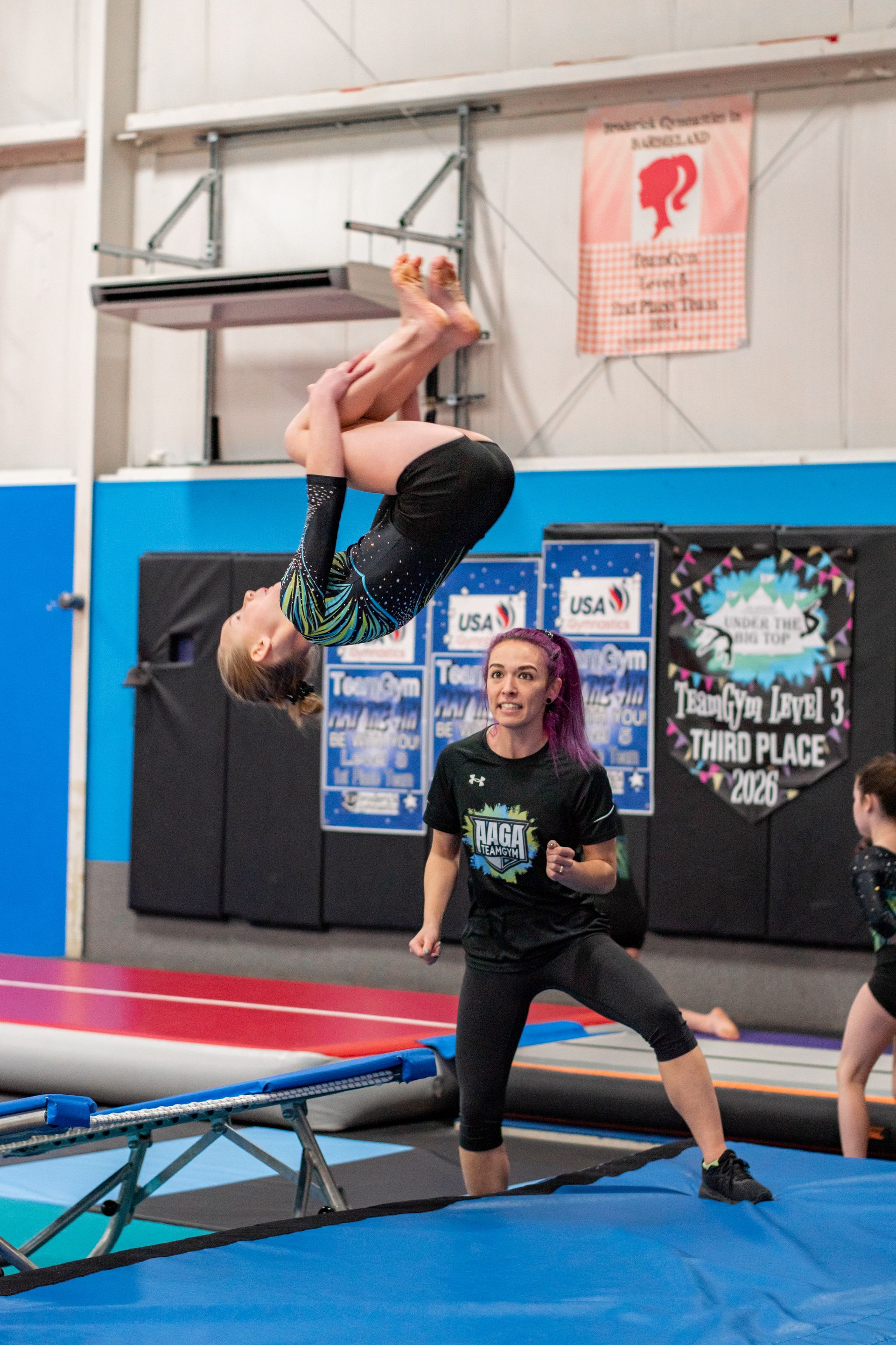 Young gymnast flipping backward above a coach on a blue mat in an indoor gym