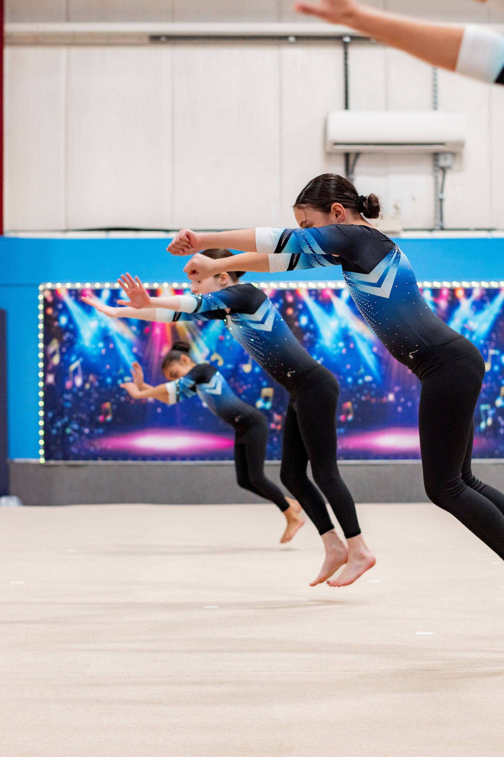 Gymnasts in blue and black leotards leap in sync on a floor exercise mat.