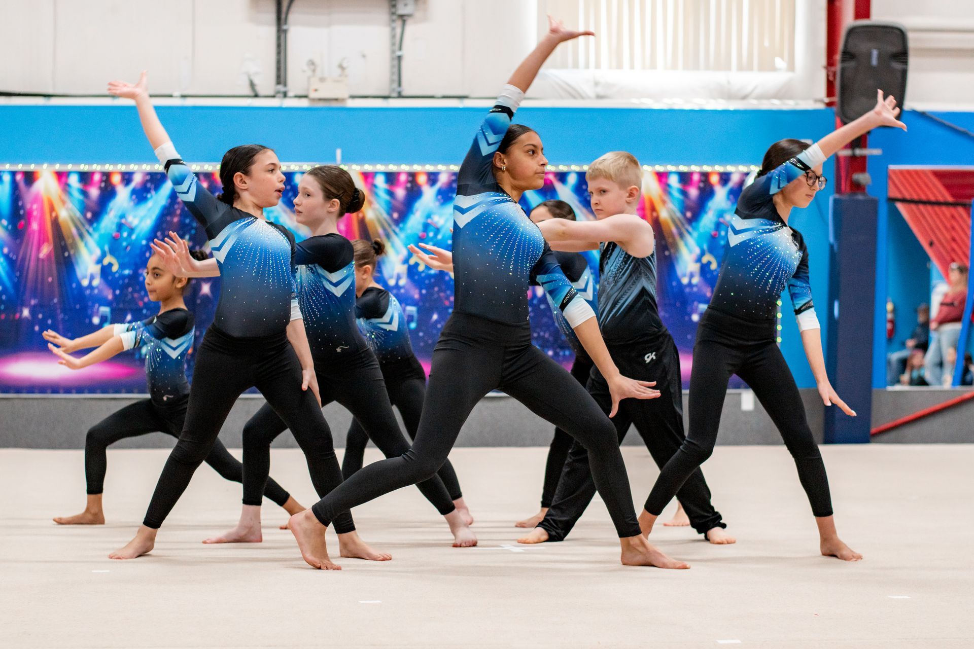 Dancers in blue costumes perform a synchronized routine in a bright studio with a colorful mural background