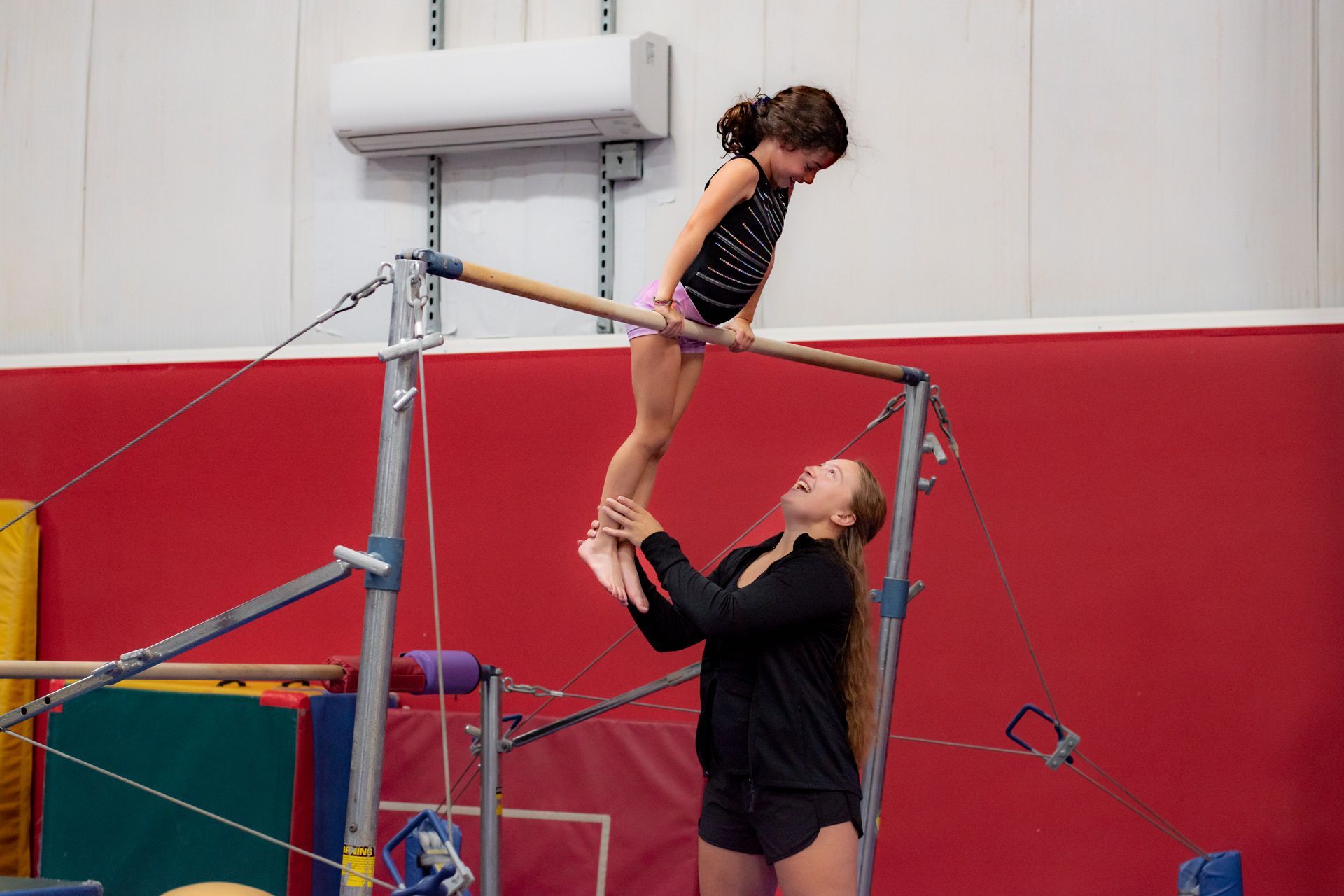 Child balancing on gymnastics bar while coach supports from below in a gym