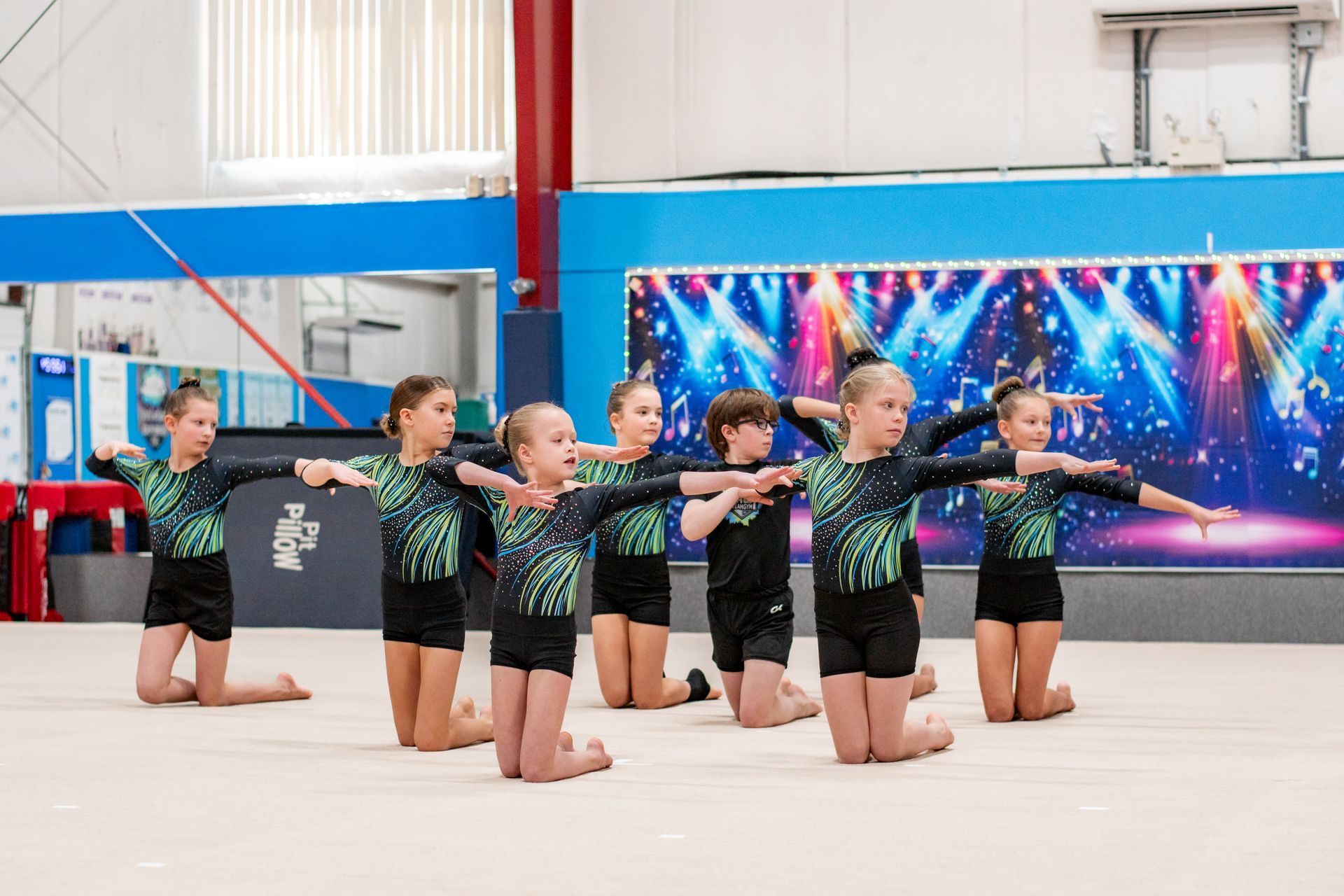 Group of young gymnasts kneeling in formation with arms extended in a gymnasium