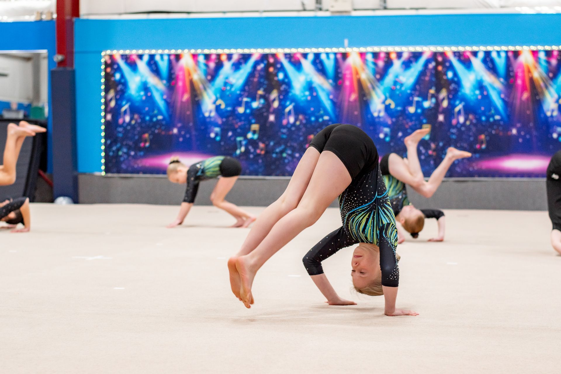 Gymnasts practicing floor routines in a bright studio, one balancing upside down in the foreground.