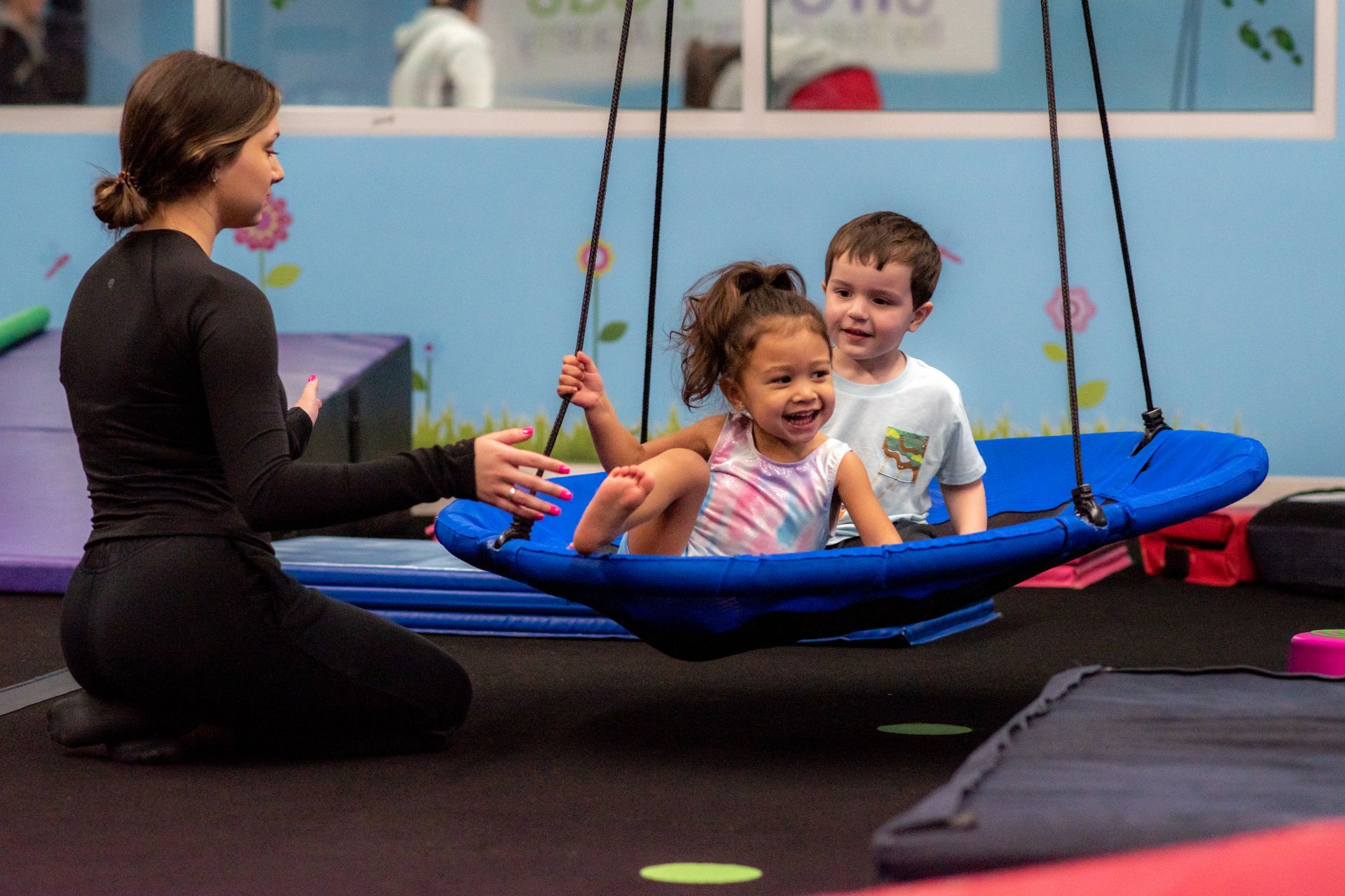 Two children on a blue swing in a gym, smiling at a kneeling instructor nearby.