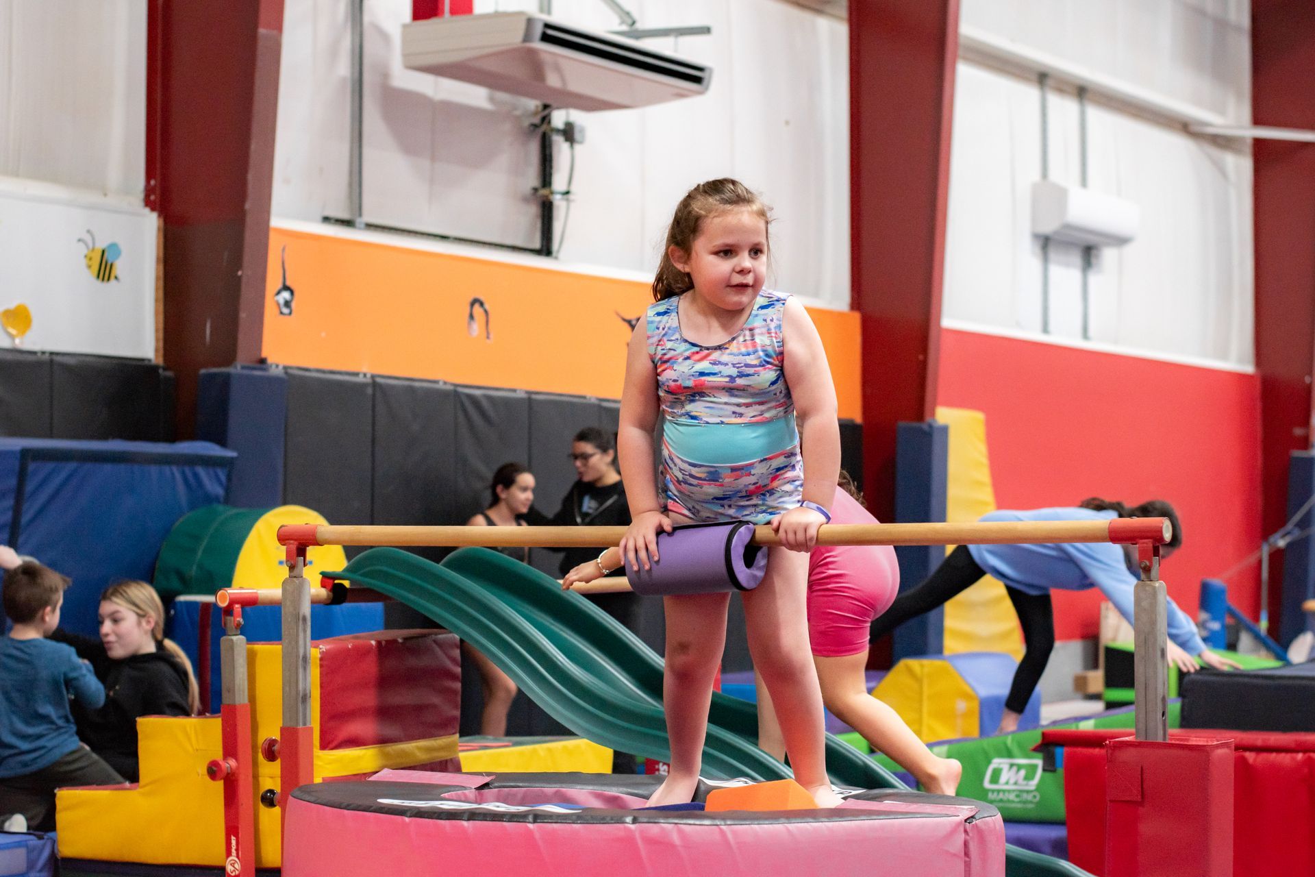 Girl on a balance beam in a colorful gym, smiling while others play in the background.