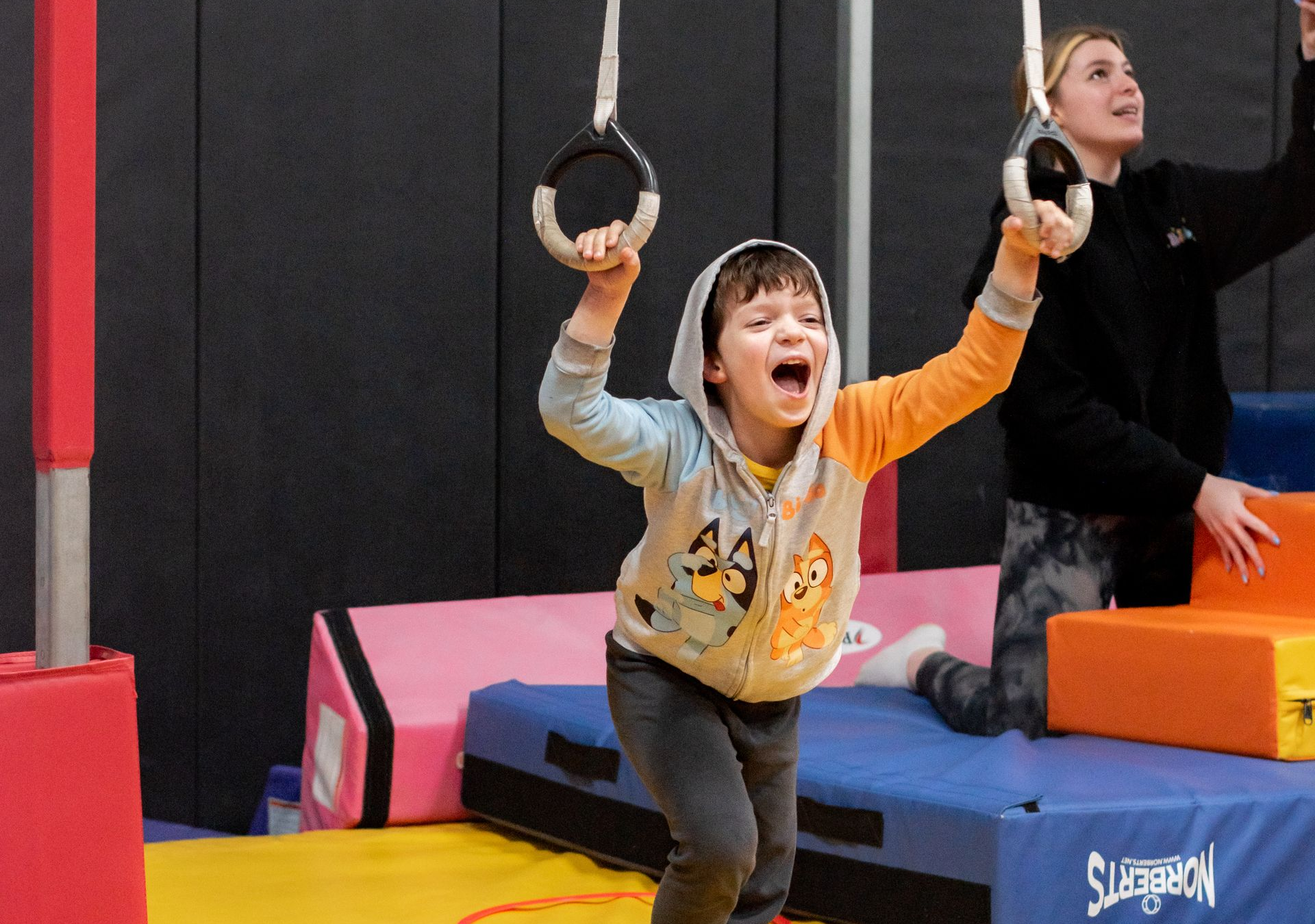 Child swinging on rings in an indoor gym, wearing a yellow shirt and smiling with mouth open.