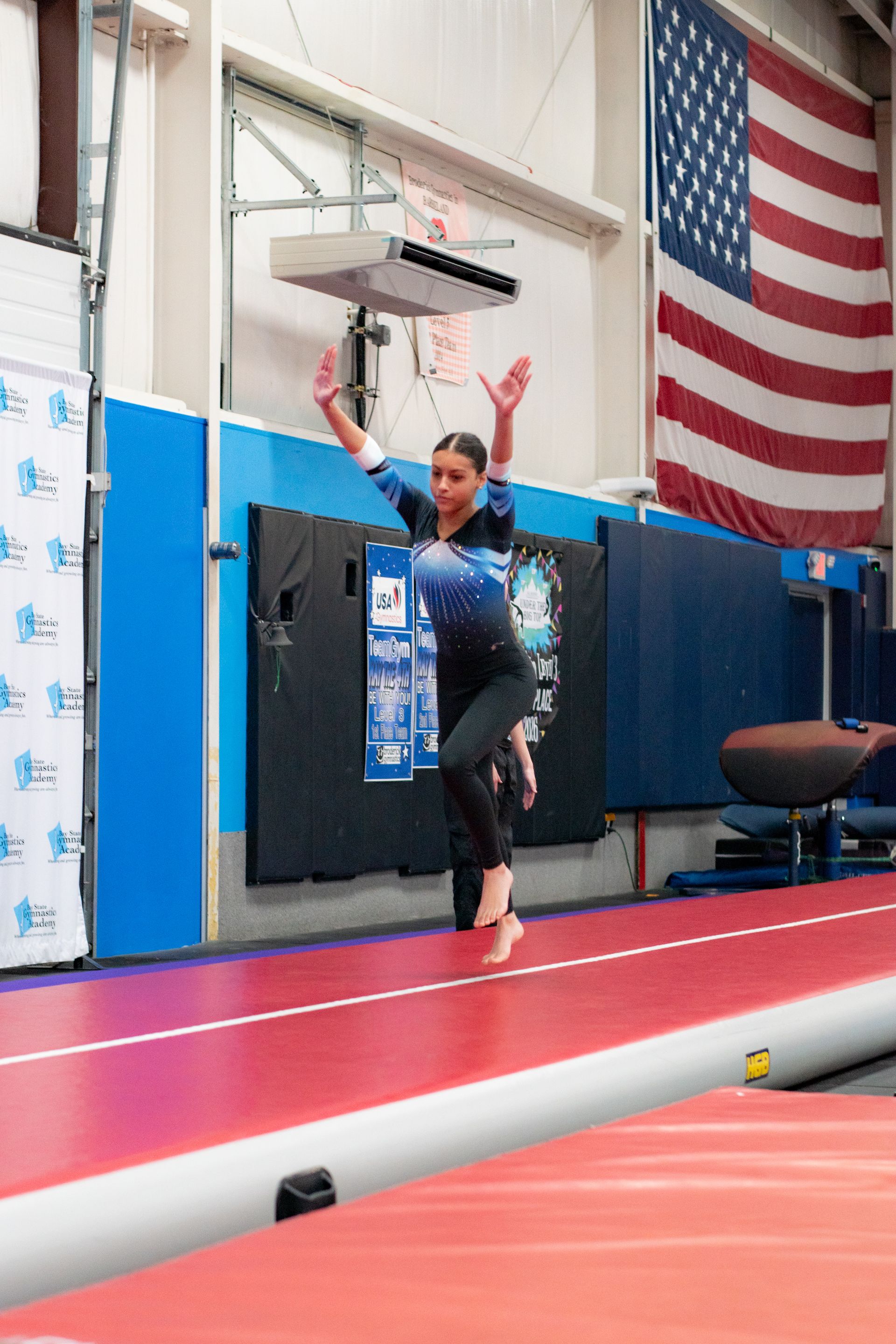 Gymnast mid-run on an indoor track with arms raised, near mats and a large American flag.