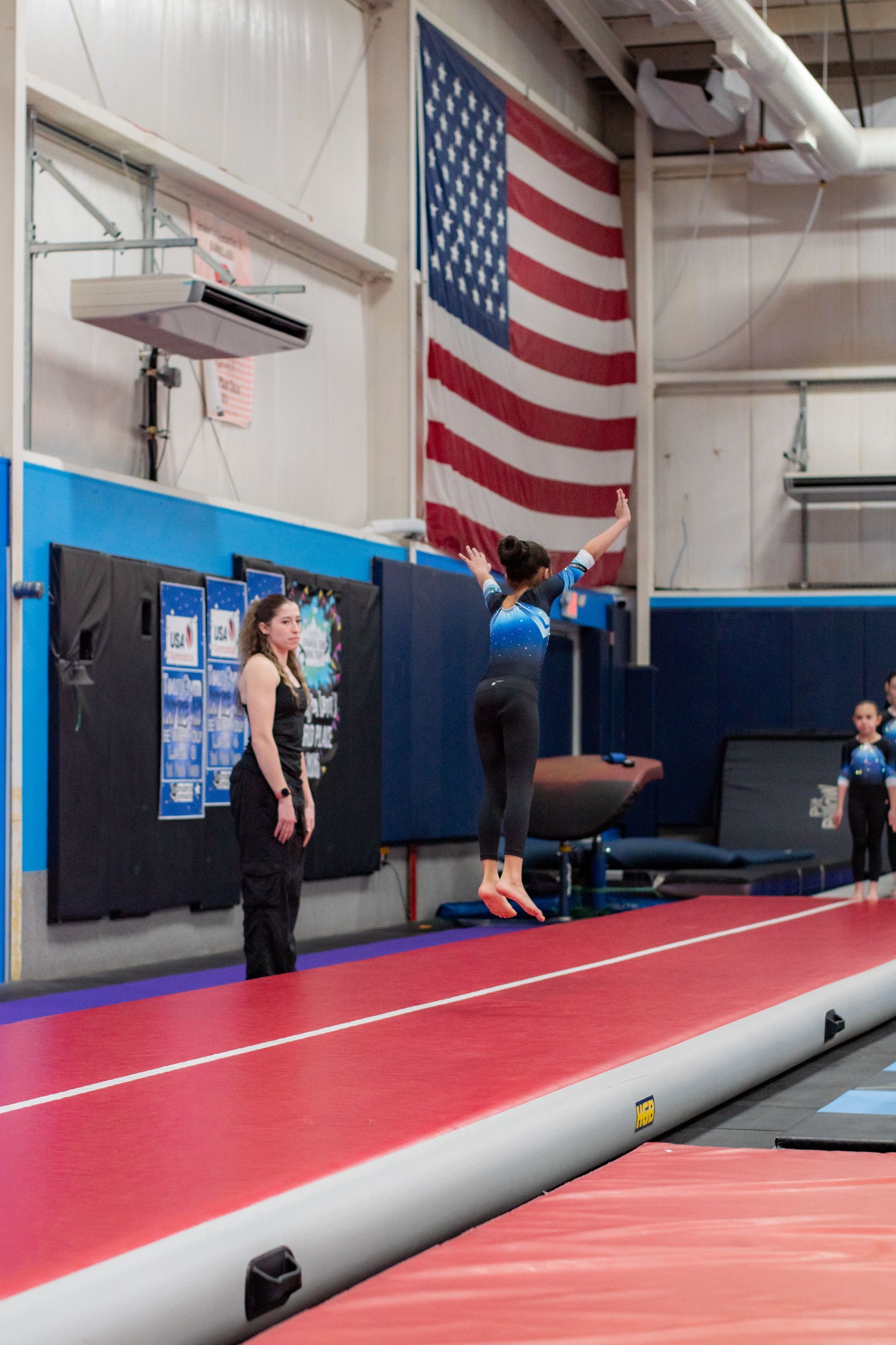 Gymnast performing a jump on a trampoline in an indoor gym with a large American flag behind her