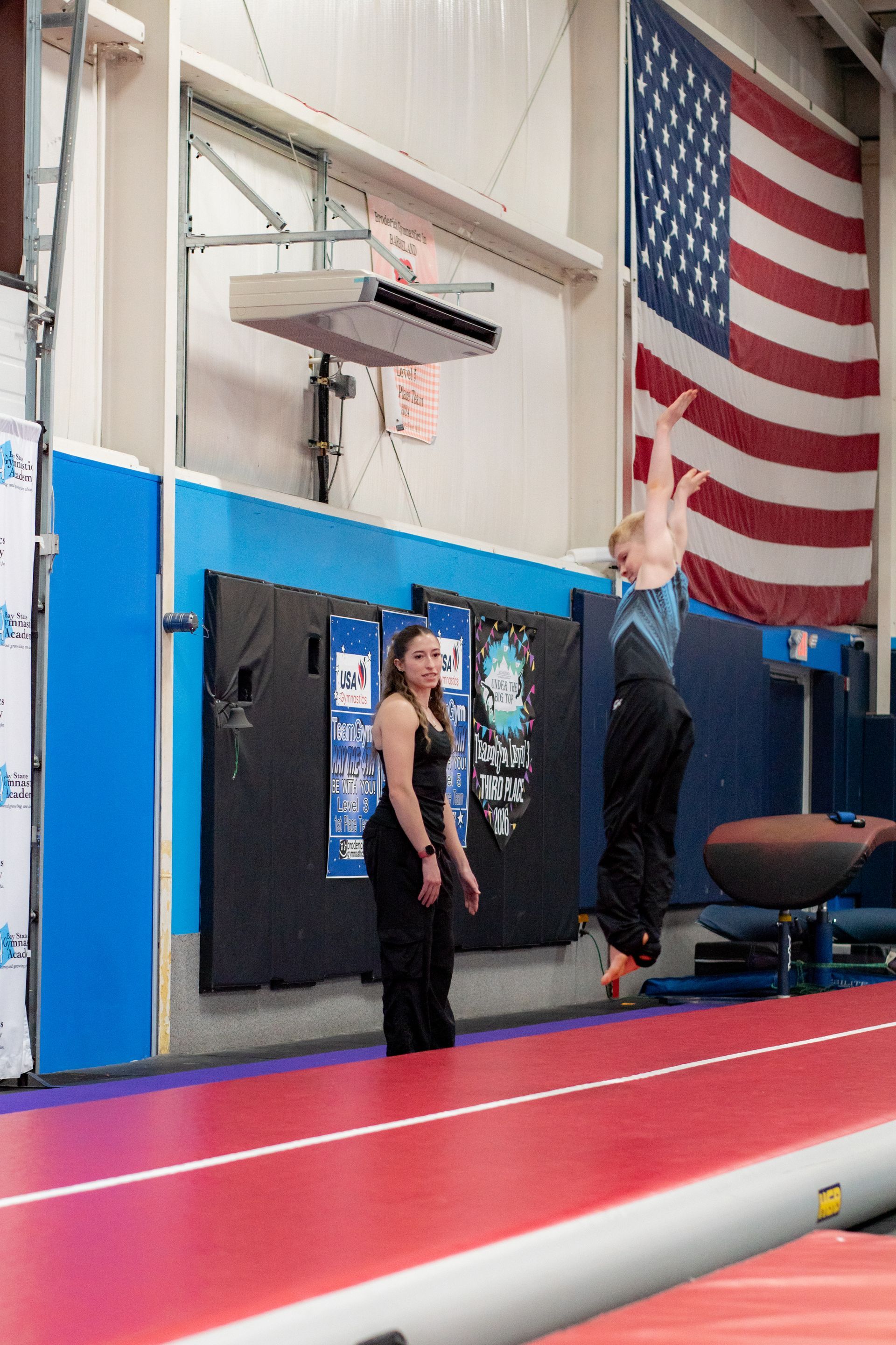 Two people in a gym, one hanging from a bar near a large American flag.
