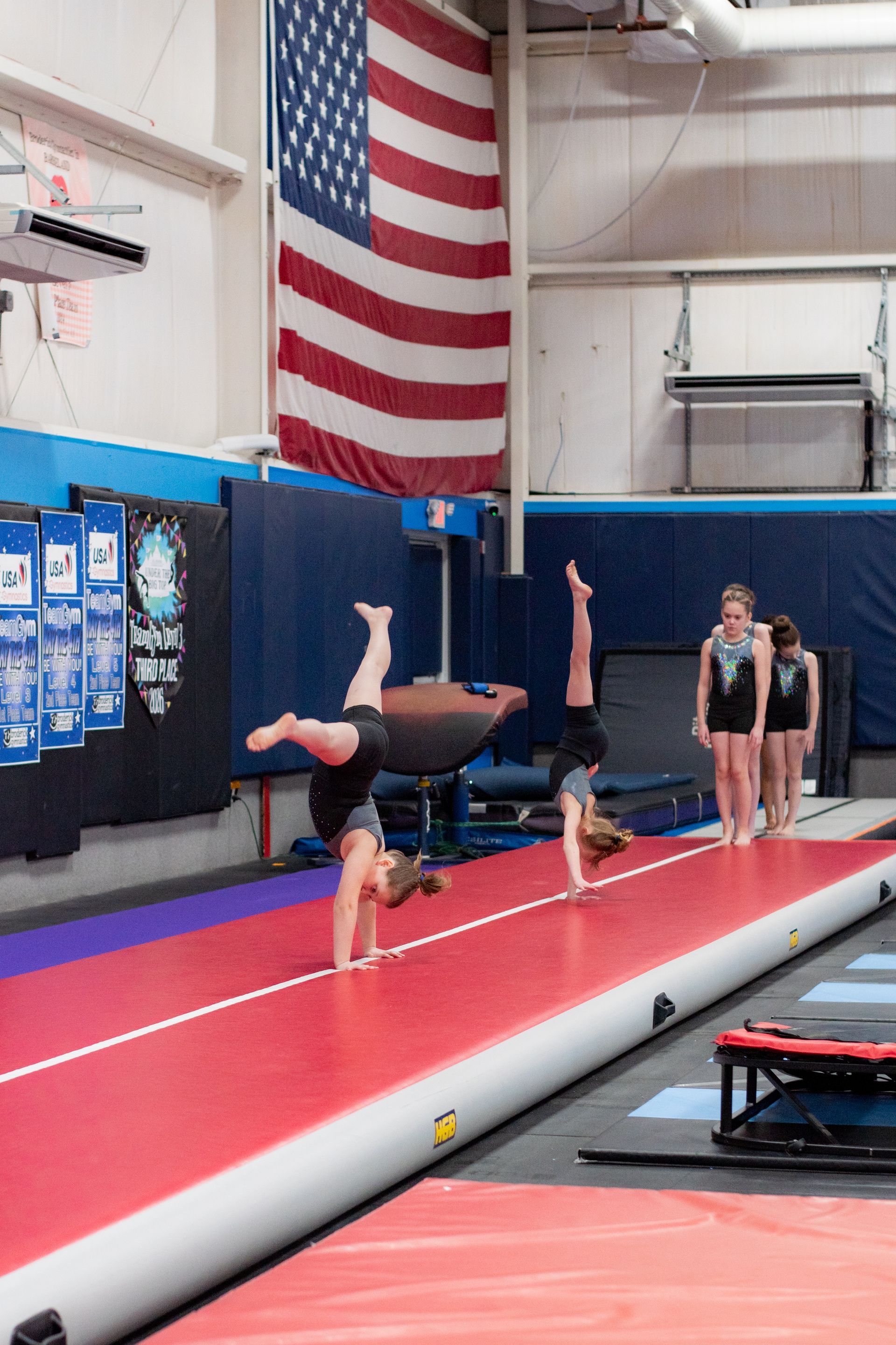 Gymnast doing a handstand on a vault runway in an indoor gym, with two athletes waiting nearby.