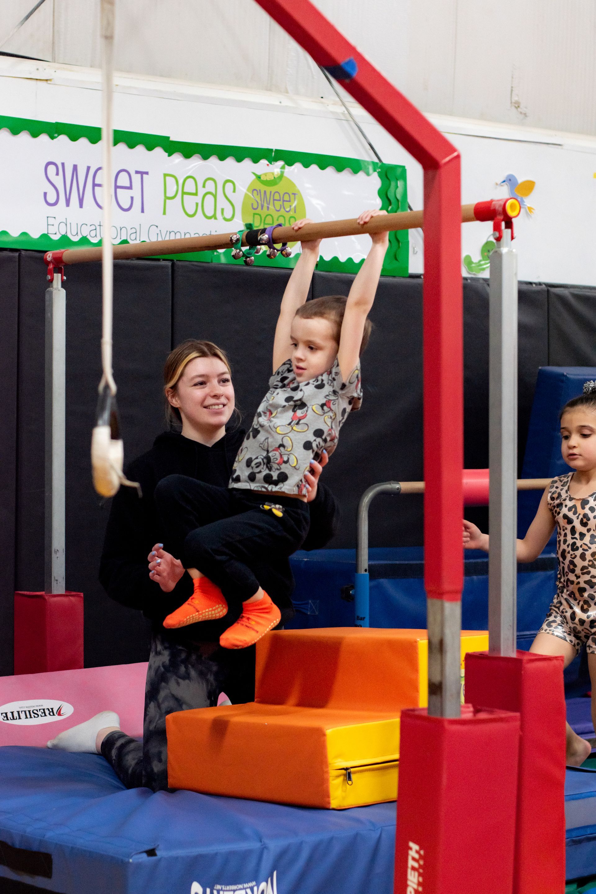 Children swinging on indoor playground bars over padded blocks, with one girl smiling and hanging from the bar