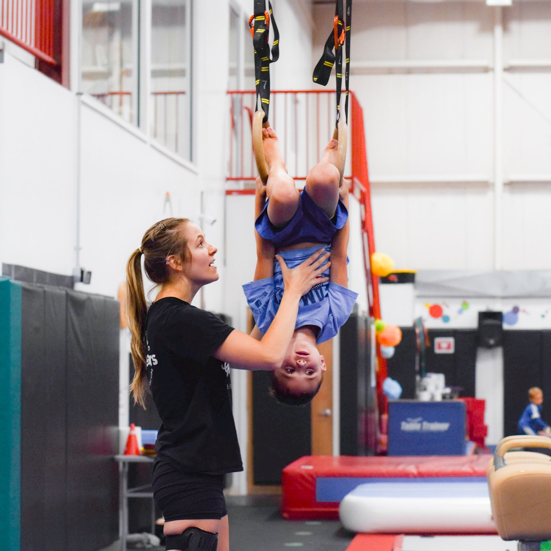 Gymnast upside down on straps while trainer spots them in an indoor gym