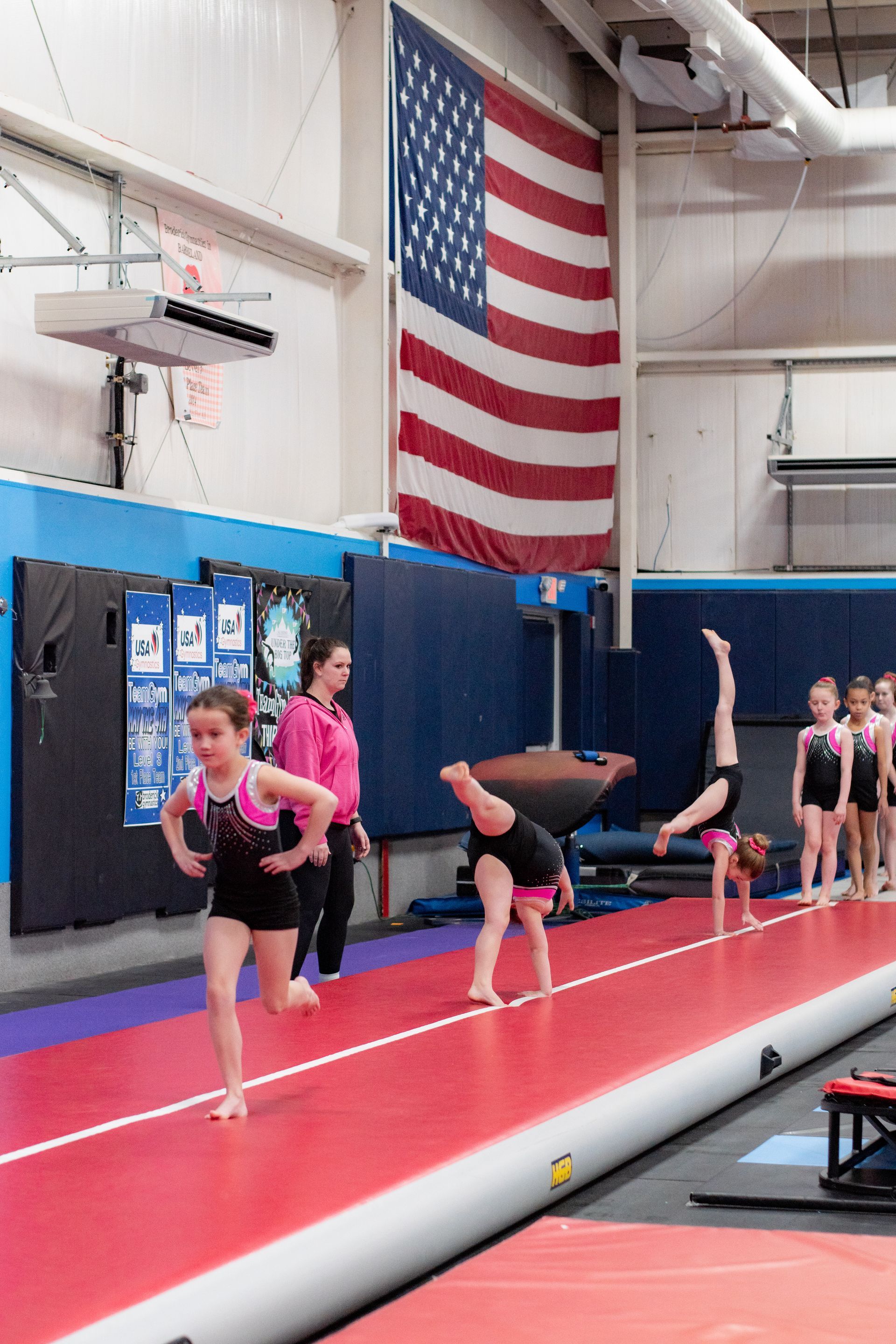 Gymnastics practice in a gym, with athletes on a red mat beneath a large American flag.