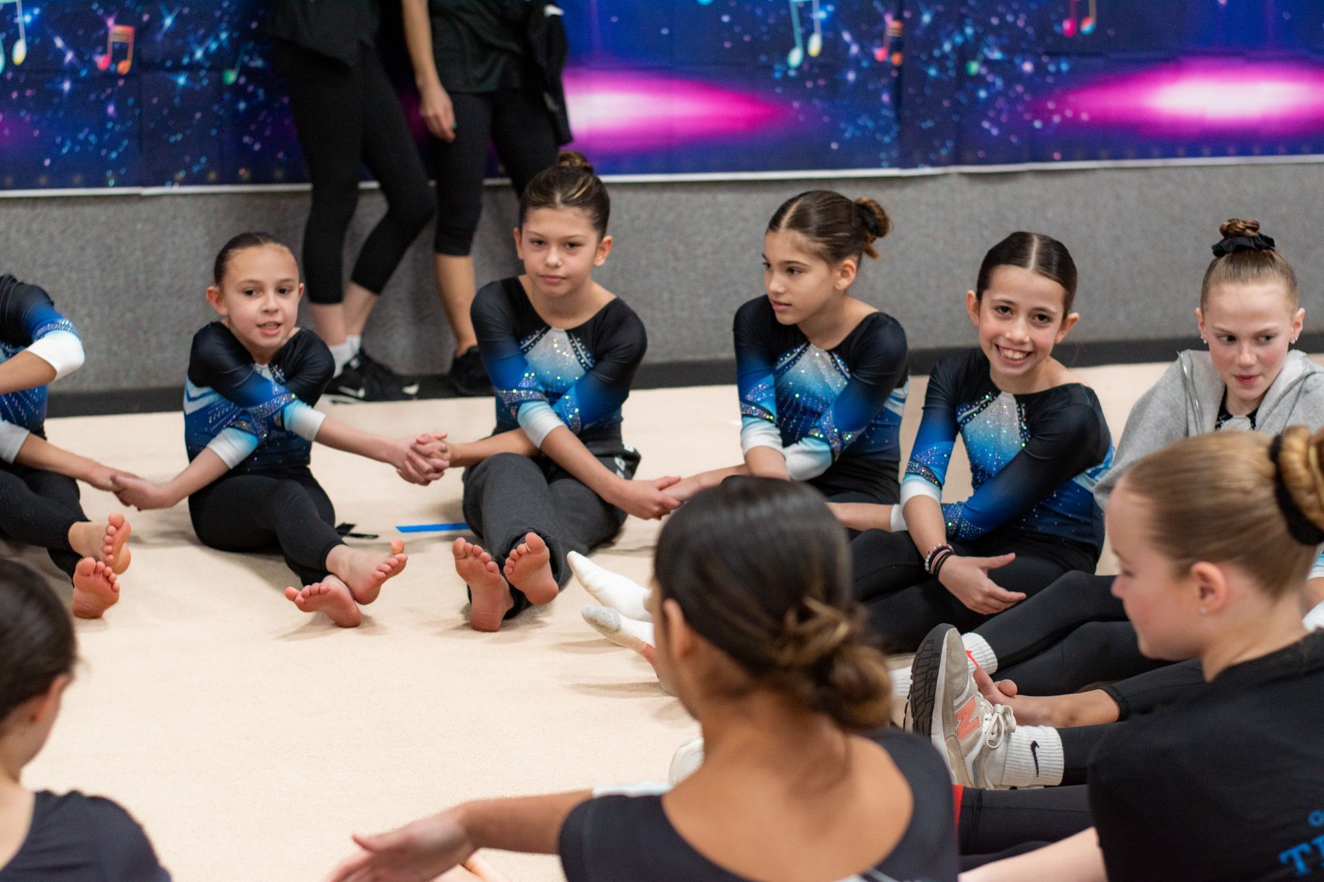 Children in blue-and-black outfits sit in a circle on a gym floor, listening together.