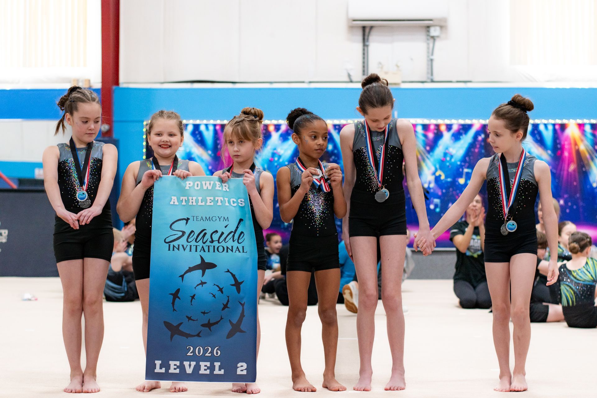 Five gymnasts in black leotards stand on a competition floor, holding a first-place trophy sign.