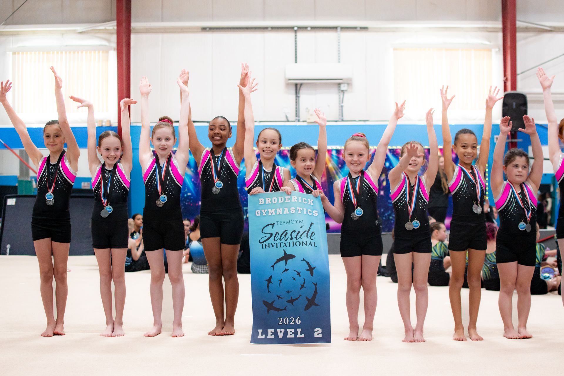 Gymnasts posing in a row with arms raised, standing behind a team sign in a gym
