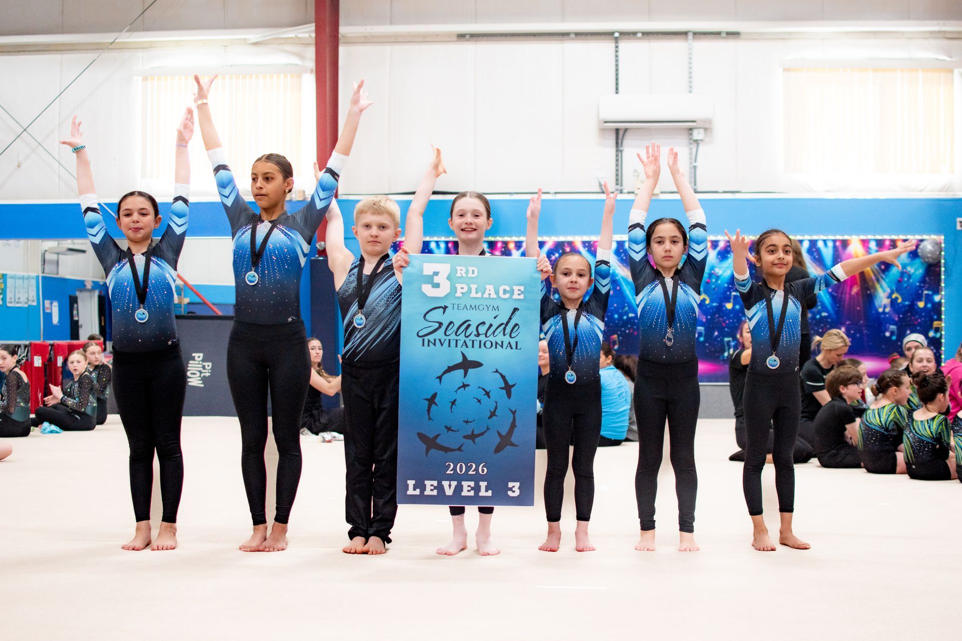 Gymnasts in blue leotards stand on a mat, raising their arms beside a competition banner.