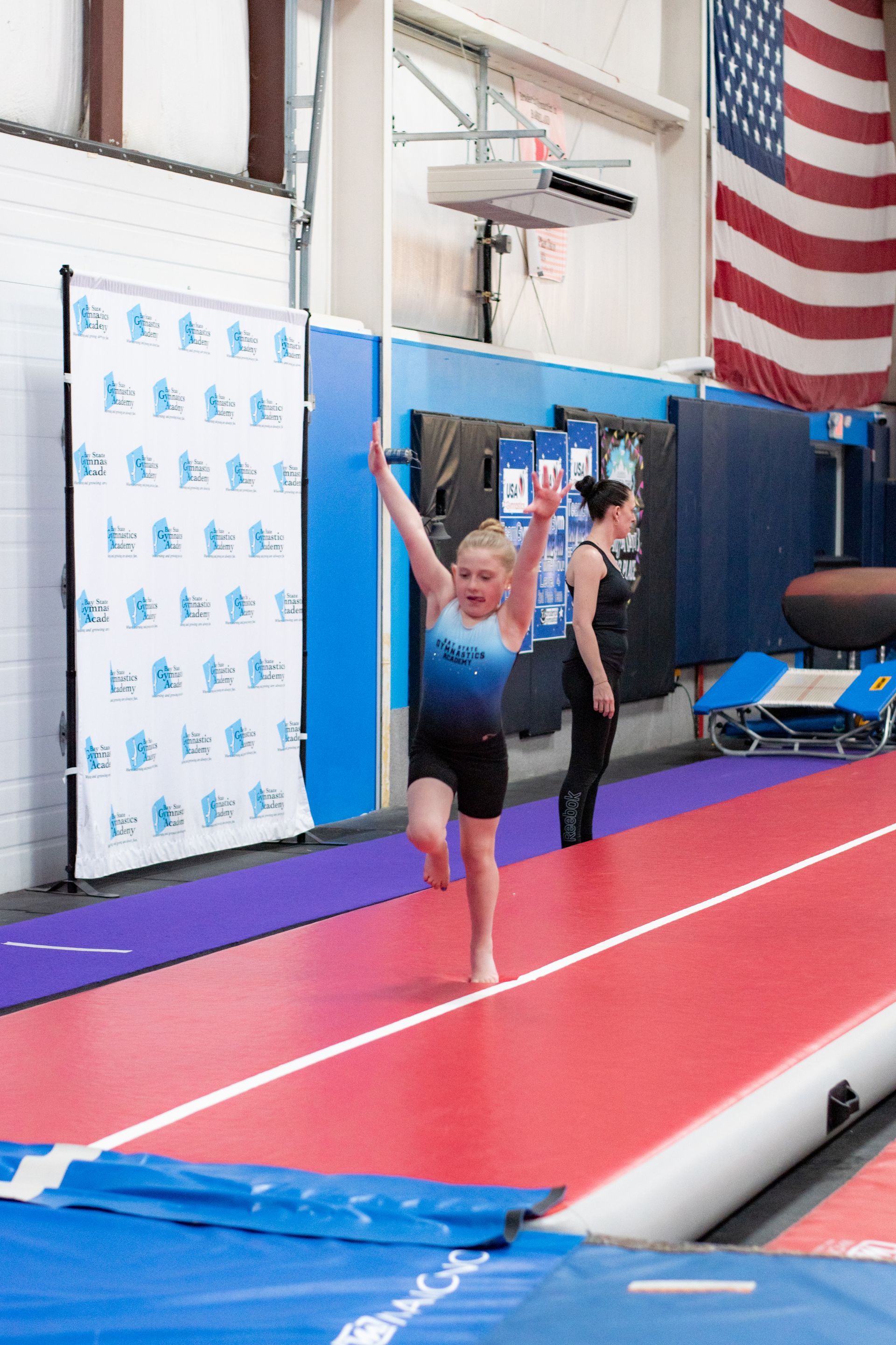 Gymnast on a red air track with arms raised, under an American flag in an indoor gym