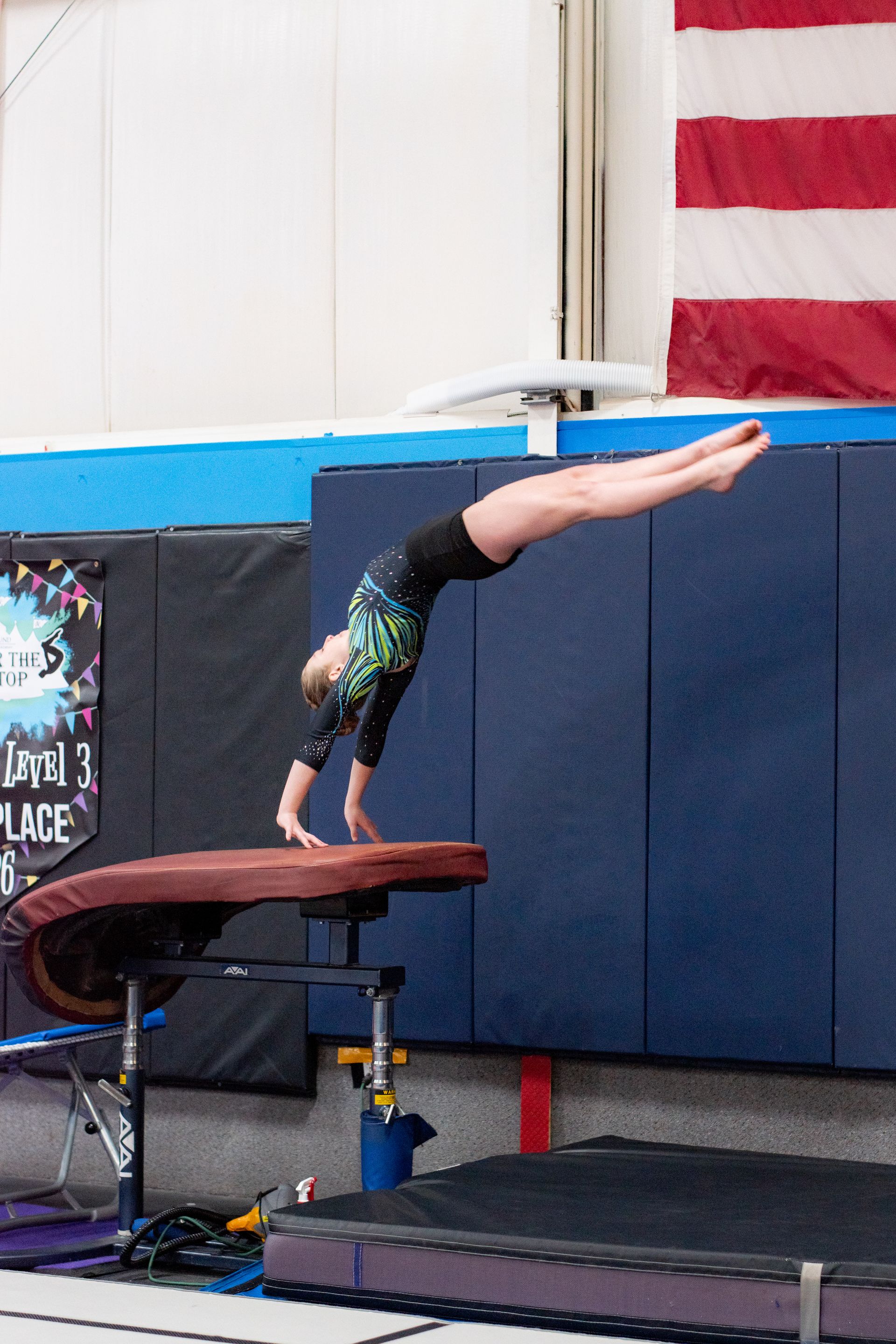 Gymnast arching backward on a balance beam in a gym, with a U.S. flag in the background