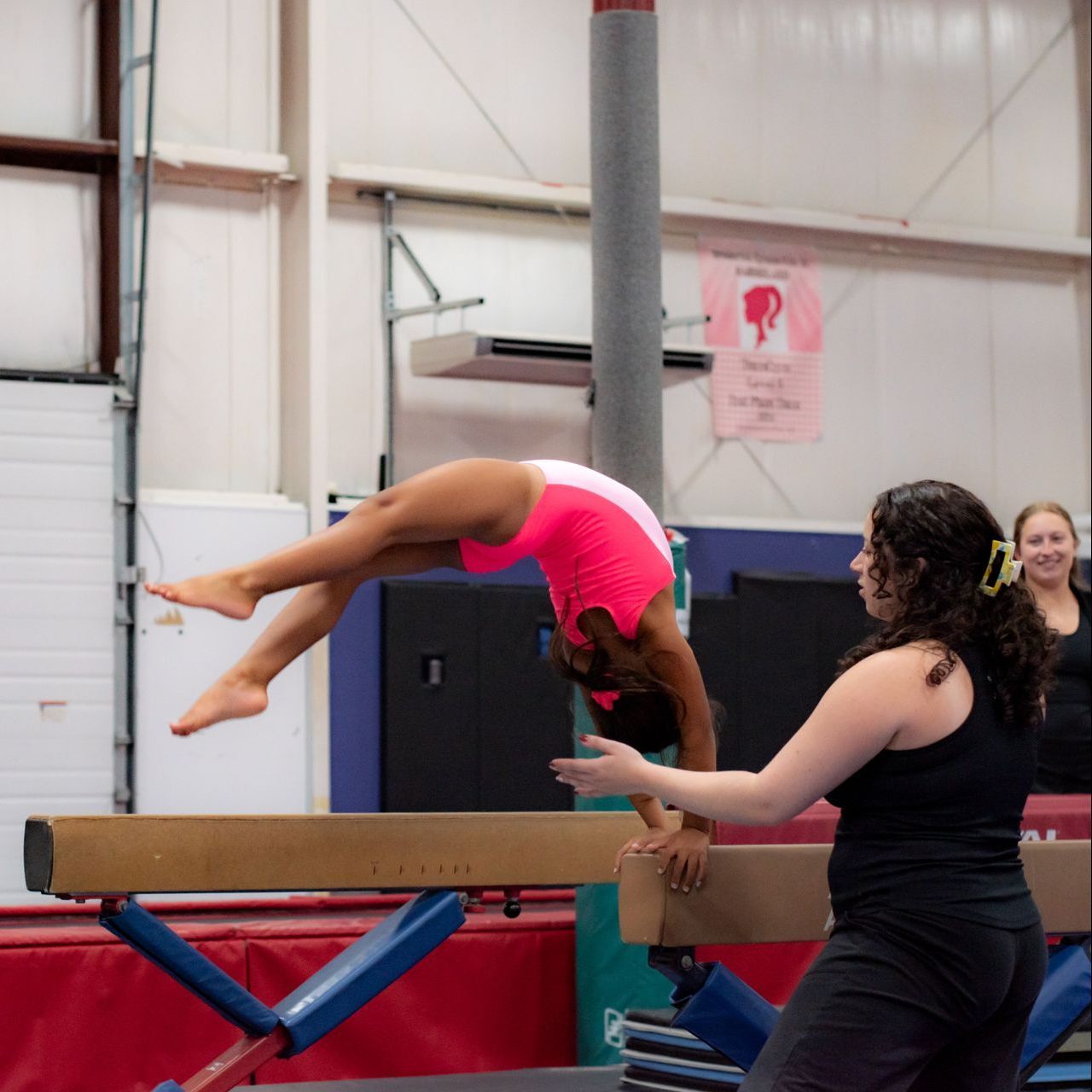 Gymnast doing a back handspring on a balance beam while a coach spots her in a training gym