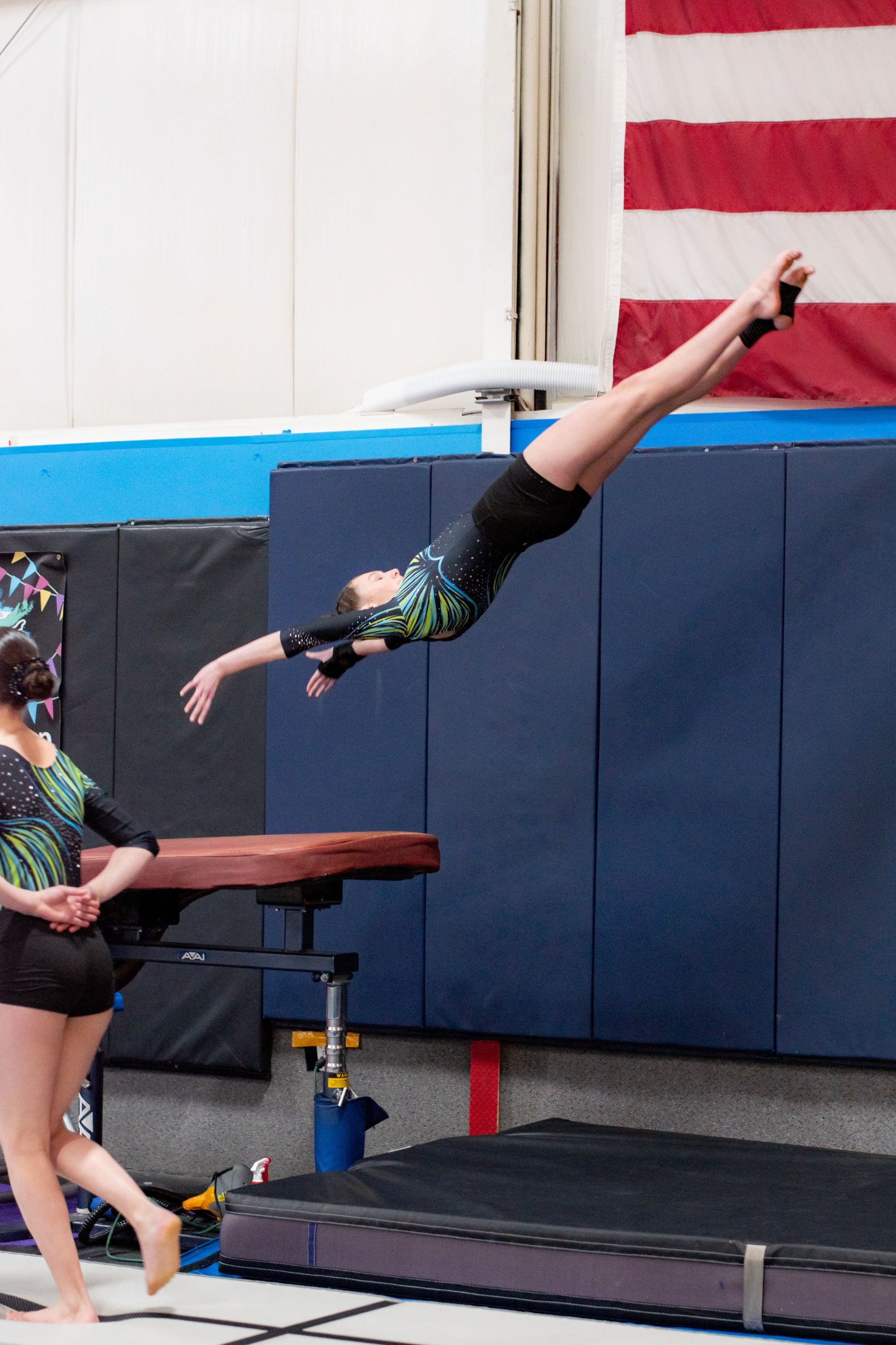Gymnast in midair vaulting over a platform in an indoor gym, with a U.S. flag on the wall.