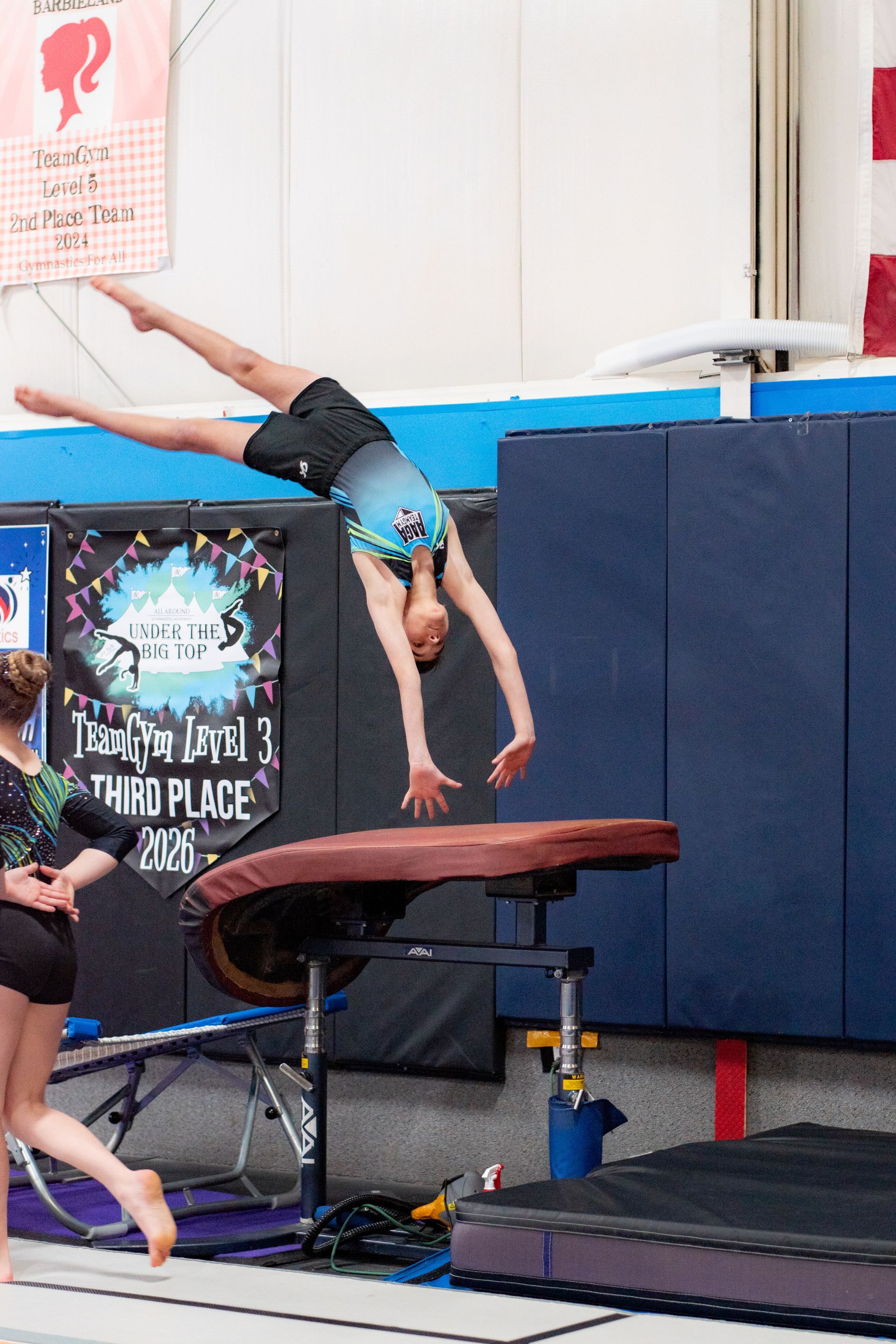 Gymnast mid-flip over a vault table in a gym, with blue padded walls and matting.