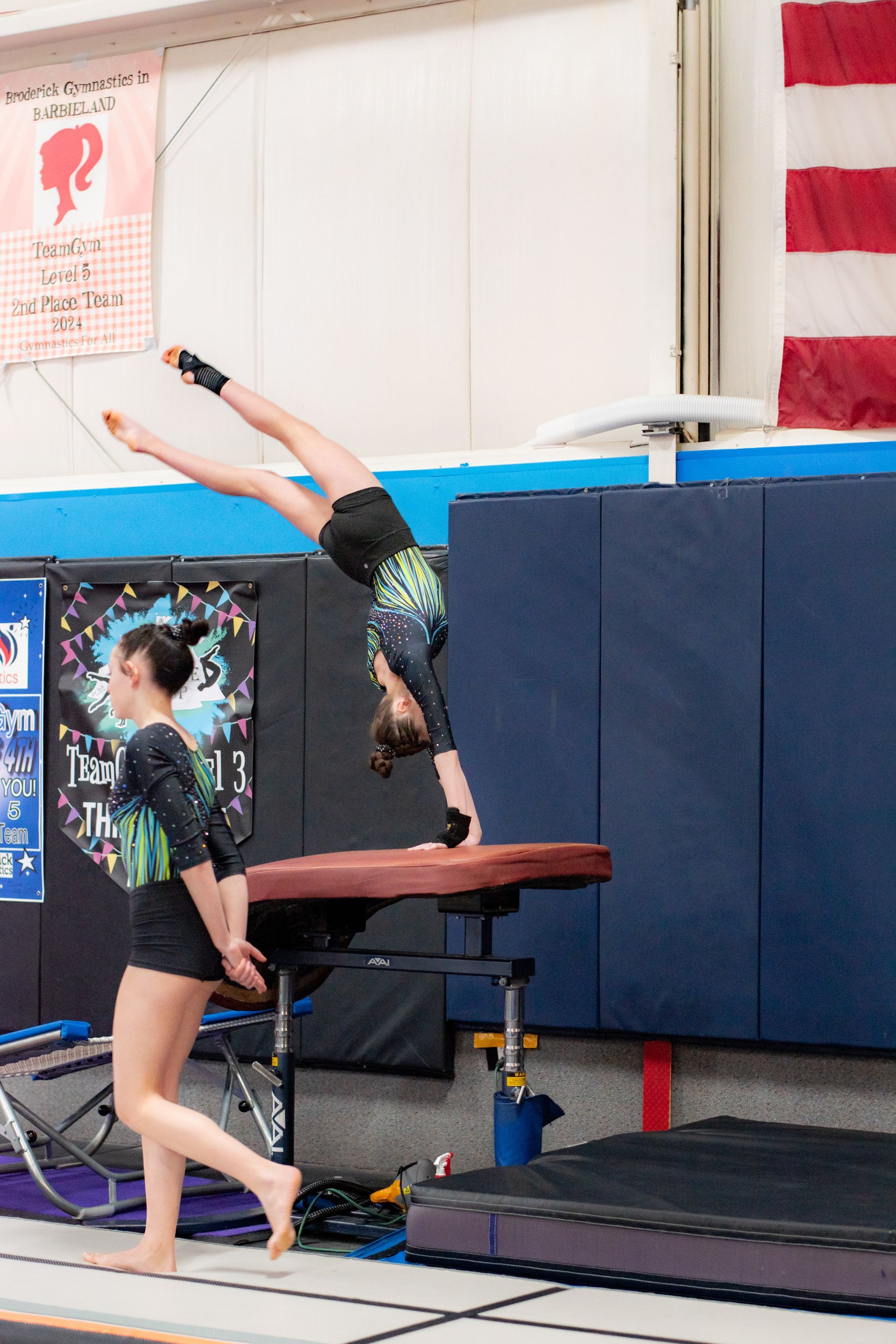 Gymnast performing a handstand on a vault table in an indoor training gym, with a coach nearby.