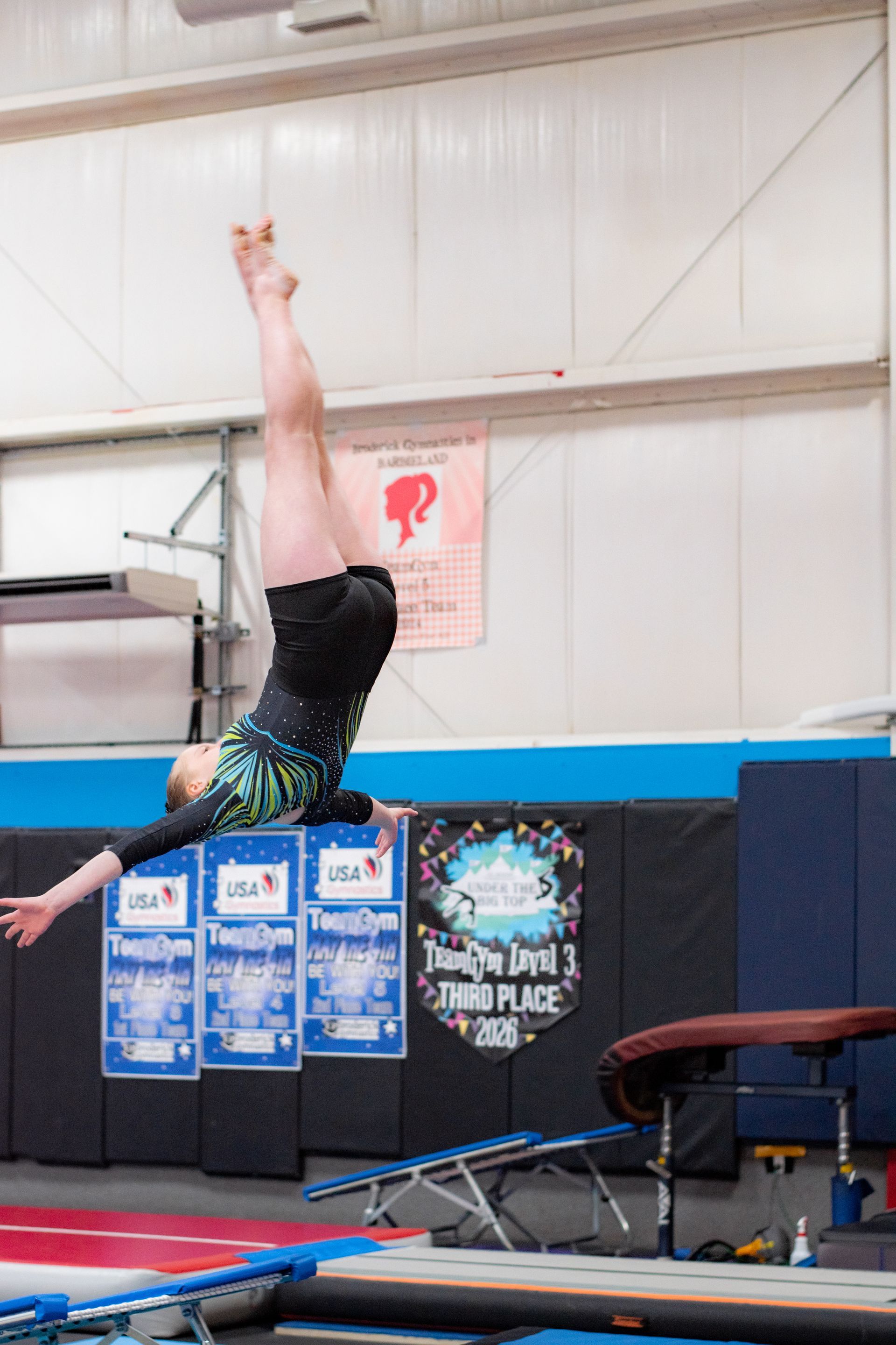 Gymnast inverted on a vault table in a gym, legs straight up, with safety mats below.
