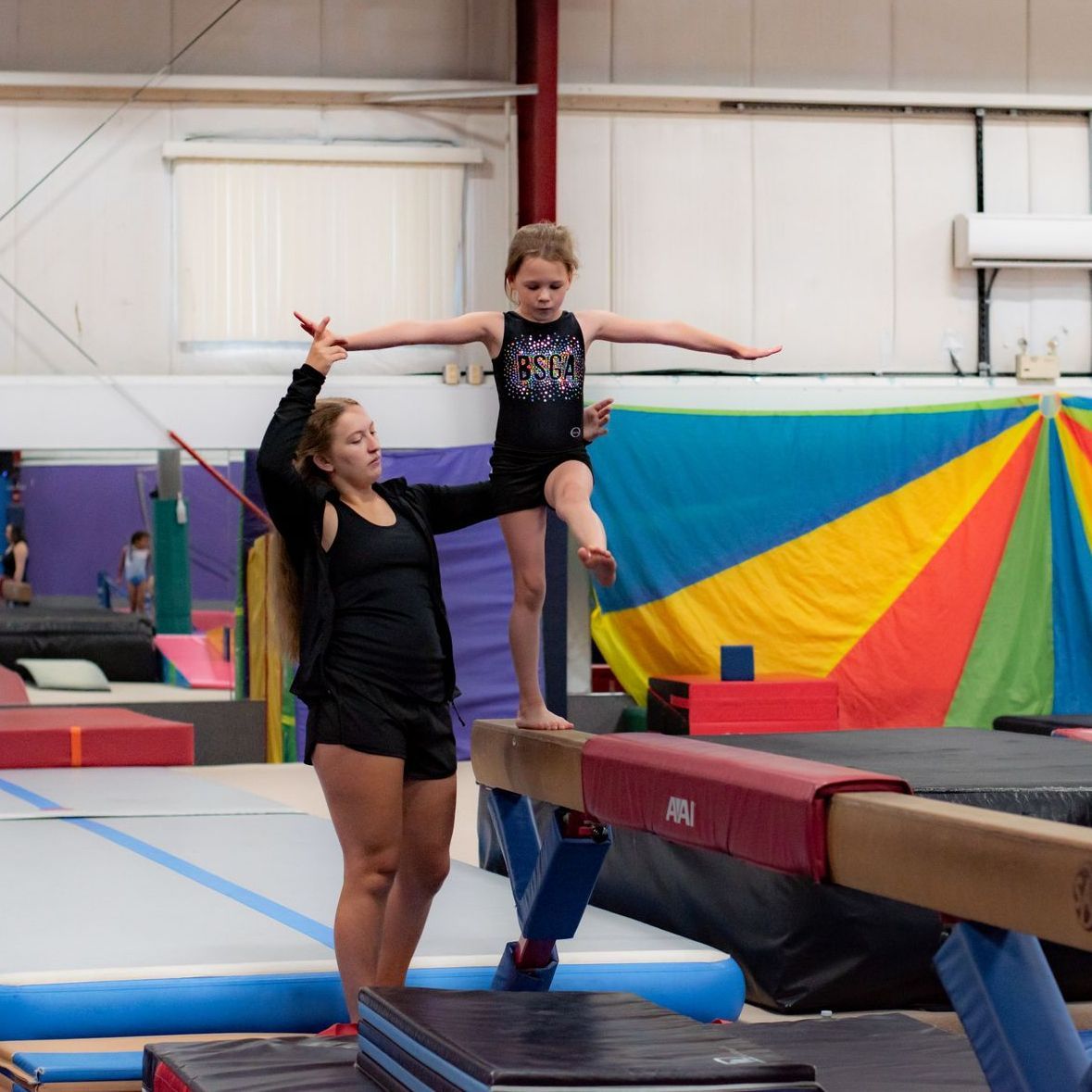 Coach spotting a child balancing on a beam in an indoor gym with colorful mats and equipment