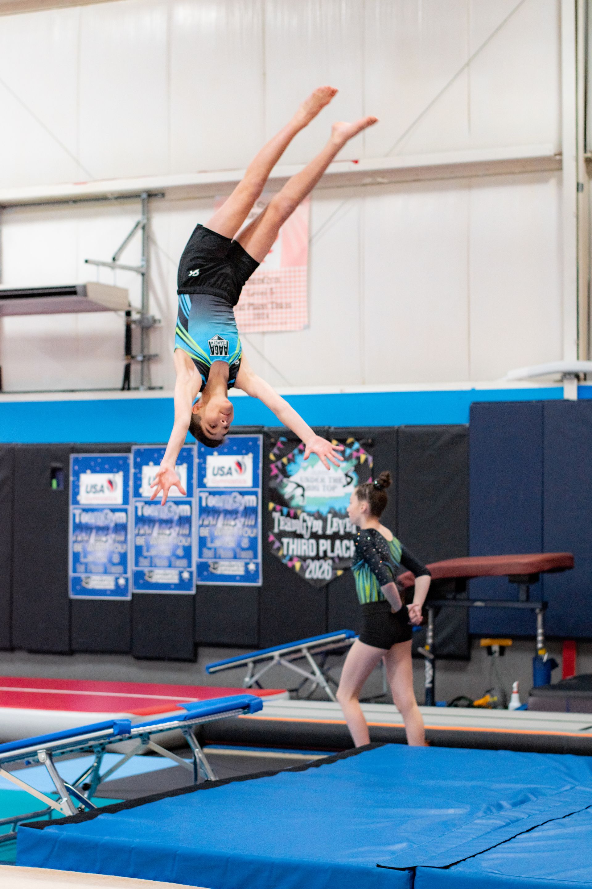 Gymnast flipping in midair over a blue mat, caught by a coach in an indoor gym.
