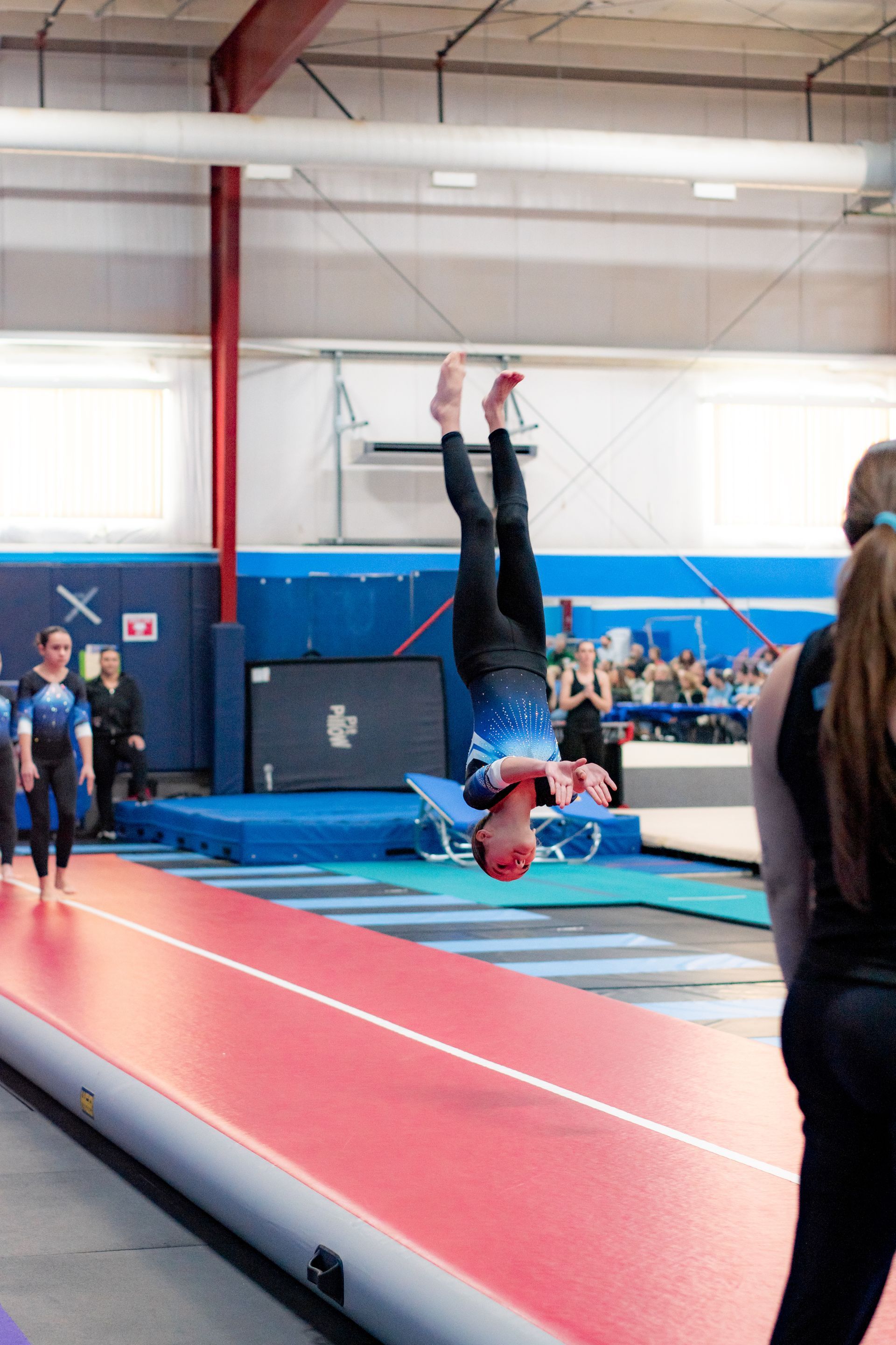 Gymnast mid-flip on a red mat in an indoor gym, with spectators in the background