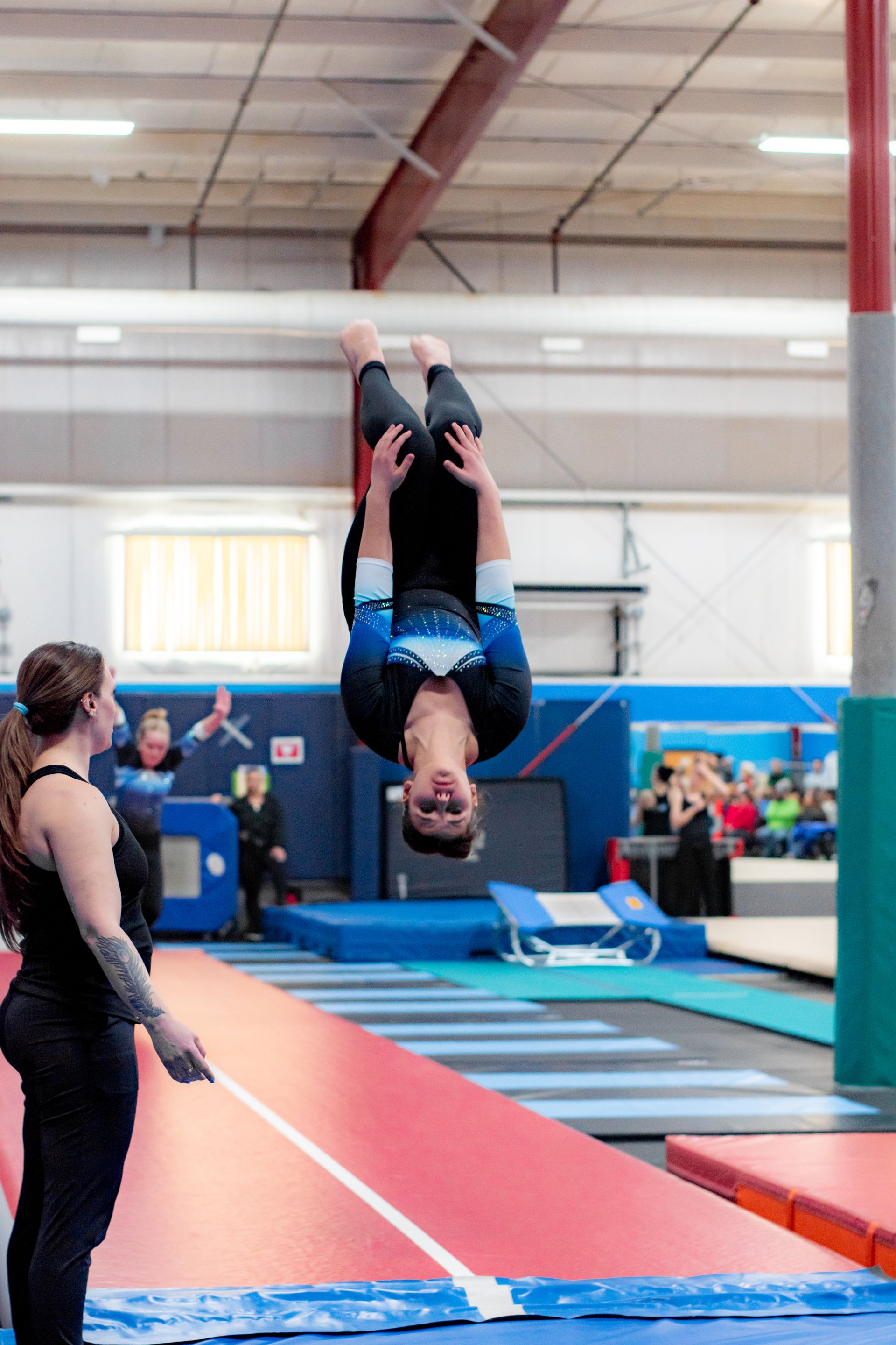 Gymnast upside down mid-flip on a gymnastics floor, with a coach watching nearby.